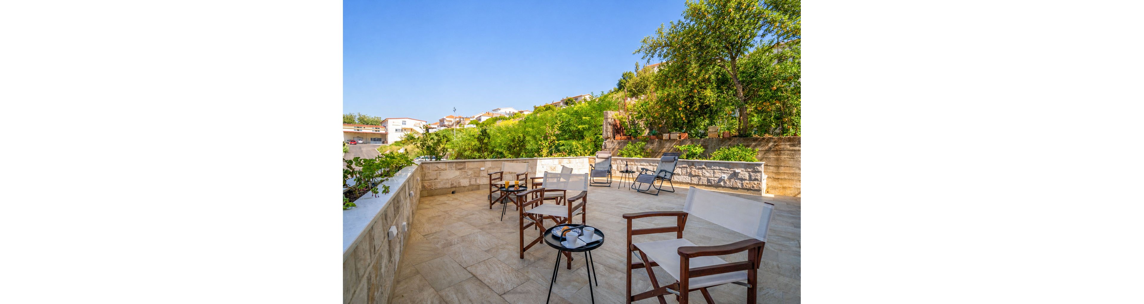 A spacious stone terrace features several wooden chairs with light fabric seats arranged around small round tables. Cups and glasses rest on the tables, suggesting a relaxed outdoor gathering. In the background, lounge chairs face the greenery and distant houses under a clear blue sky. Lush trees and shrubs surround the terrace, creating a peaceful and inviting atmosphere.