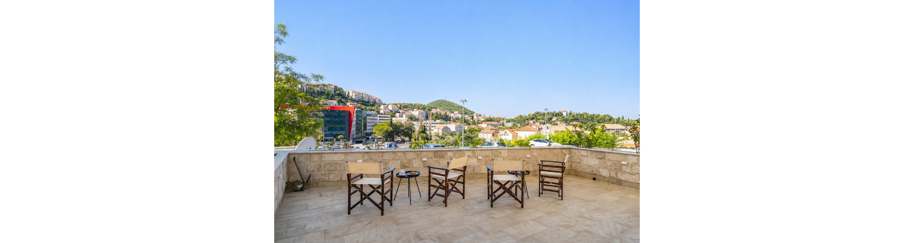 A spacious stone terrace features several wooden chairs and small round tables arranged for outdoor seating. The terrace overlooks a hillside cityscape with residential buildings, trees, and a few modern structures in the distance. Clear blue skies and bright sunlight create a warm, inviting atmosphere. The scene suggests a relaxed setting for conversation or enjoying the view.