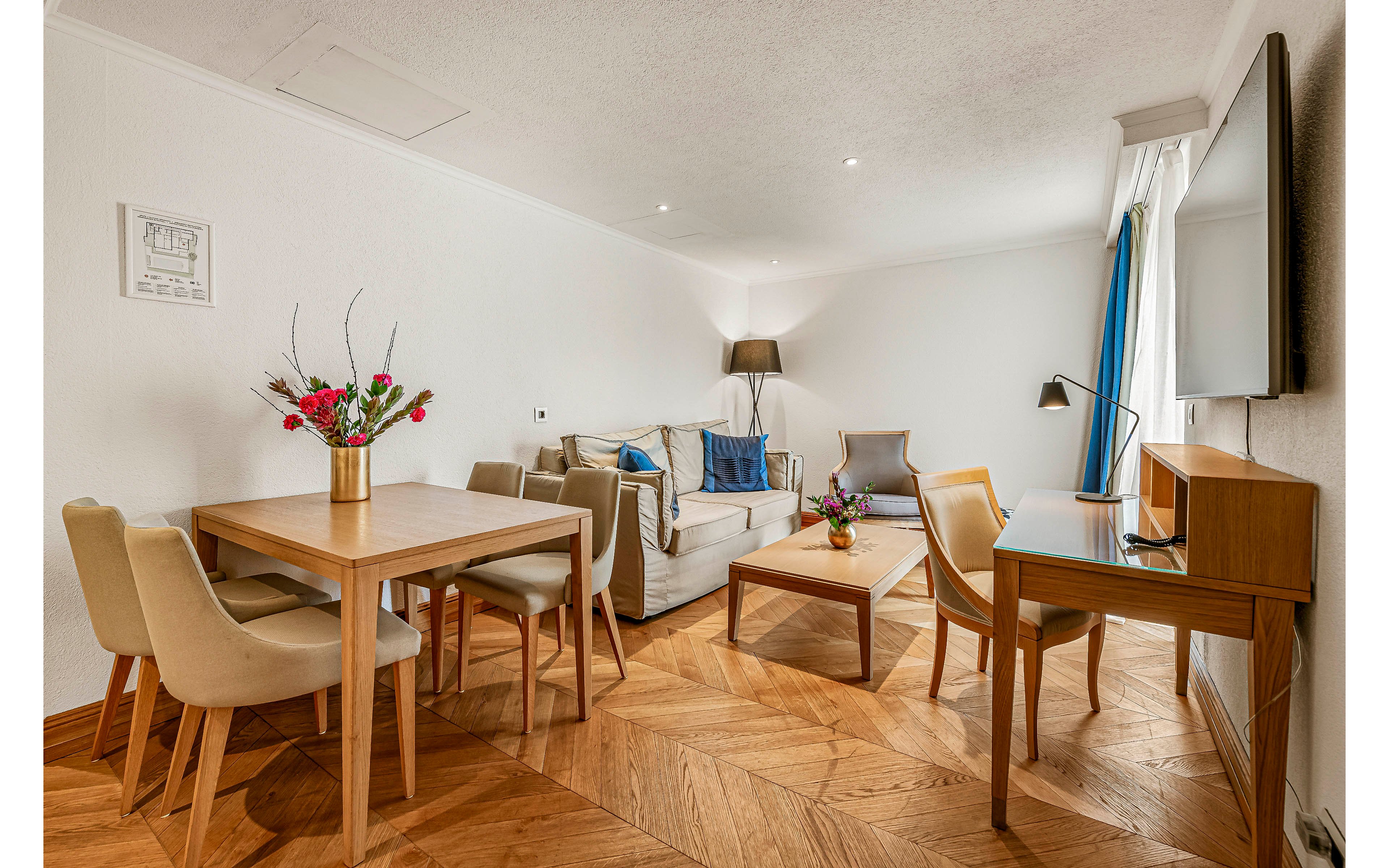 A contemporary living room features a neutral sofa, wooden dining table with chairs, and a coffee table adorned with a floral arrangement. The space is well-lit with natural light from a window, complemented by a floor lamp and desk lamp. The room has a warm wood floor, minimalist decor, and a wall-mounted television, creating a cozy and inviting atmosphere.