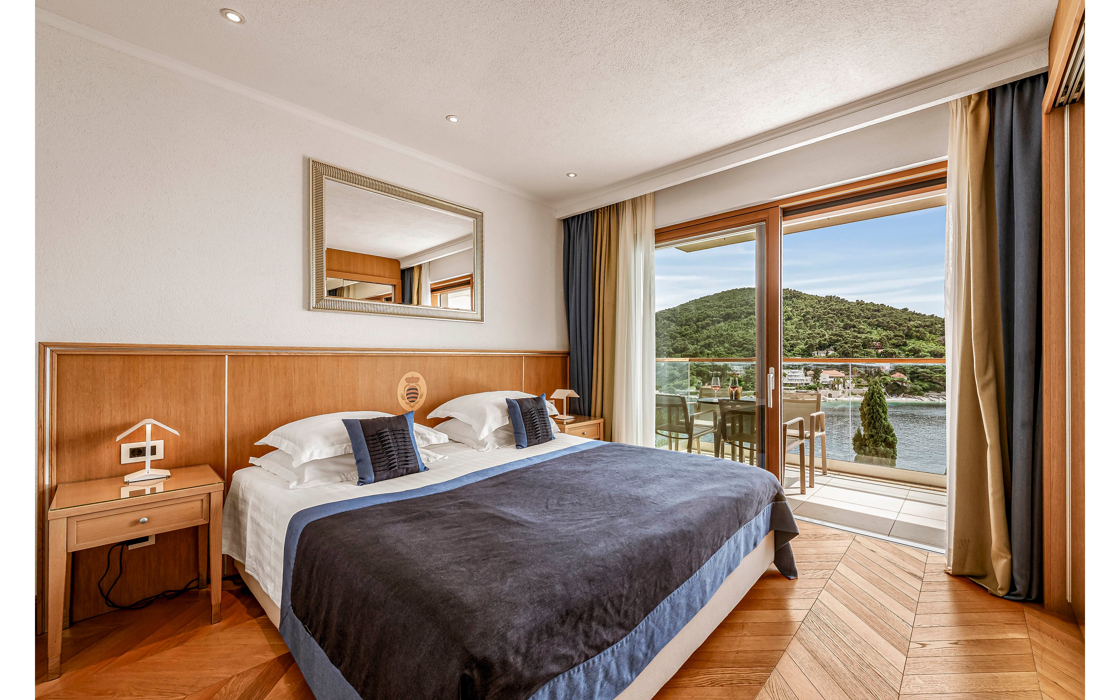 A contemporary hotel room features a large bed with navy and white linens, flanked by wooden nightstands. The space is bright, with natural light streaming in from a glass door that opens onto a balcony with outdoor seating. The balcony offers a scenic view of a lake and green hills, creating a tranquil atmosphere. The room is decorated in neutral tones with blue accents and geometric wood flooring.