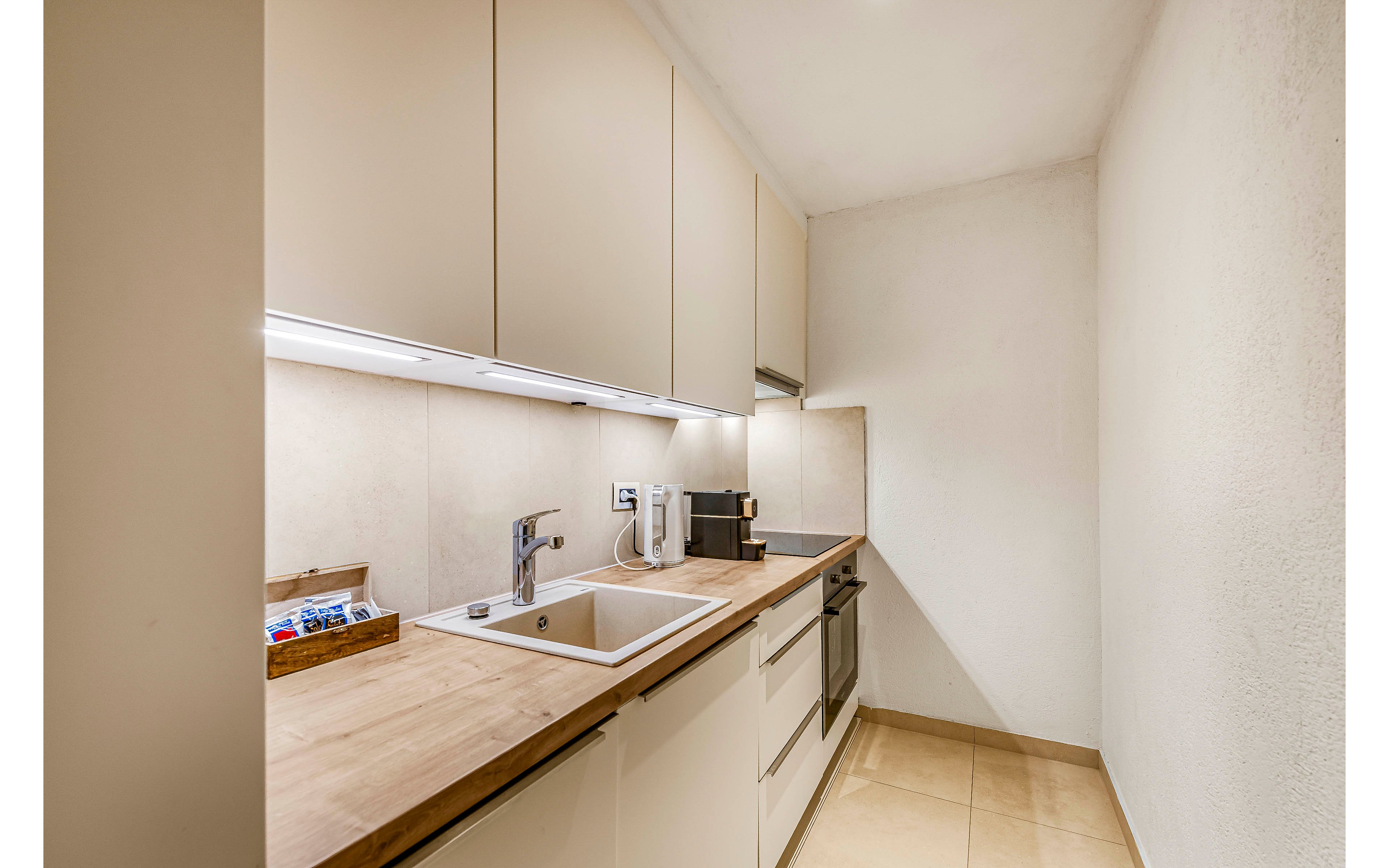 A compact, contemporary kitchen features sleek beige cabinetry and a wood countertop. The space includes a stainless steel sink, faucet, and built-in appliances, with a coffee machine and utensil holder visible. The setting is clean and well-lit, with neutral tones and a minimalist style. No visible text or numeric values are present in the image.