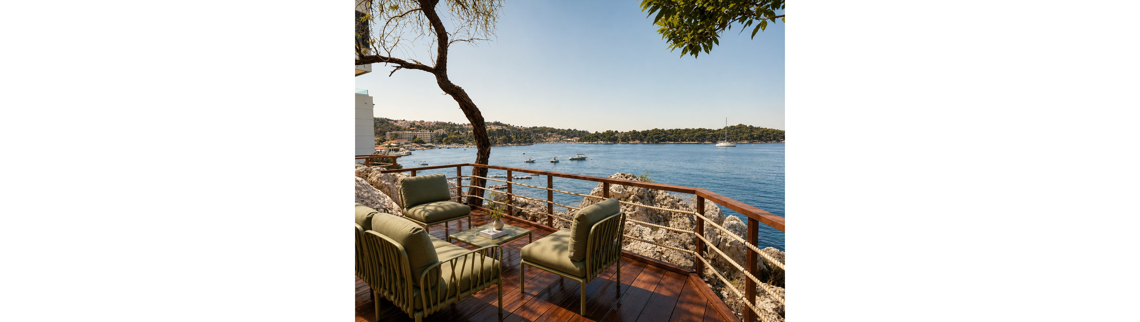 An outdoor terrace with cushioned lounge chairs and a small table overlooks a calm blue bay. The wooden deck is bordered by a rope and metal railing, set above rocky shoreline. Boats and a distant sailboat float on the water, with a coastal town visible across the bay. Warm sunlight and clear skies create a relaxed, vacation-like atmosphere.