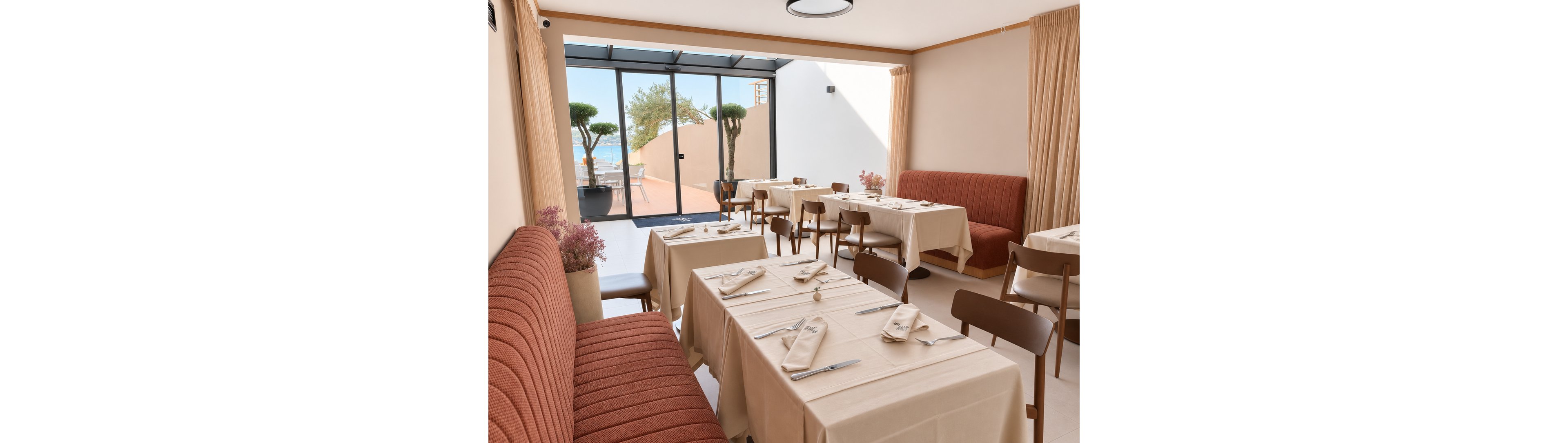 A contemporary restaurant dining room features neatly arranged tables covered with light beige tablecloths and matching napkins. Upholstered banquette seating in a warm rust tone lines one side, complemented by simple wooden chairs. Large glass doors open to a terrace with potted trees and a view of the sea in the background. Soft natural light and neutral curtains create an airy, relaxed atmosphere suitable for casual or fine dining.