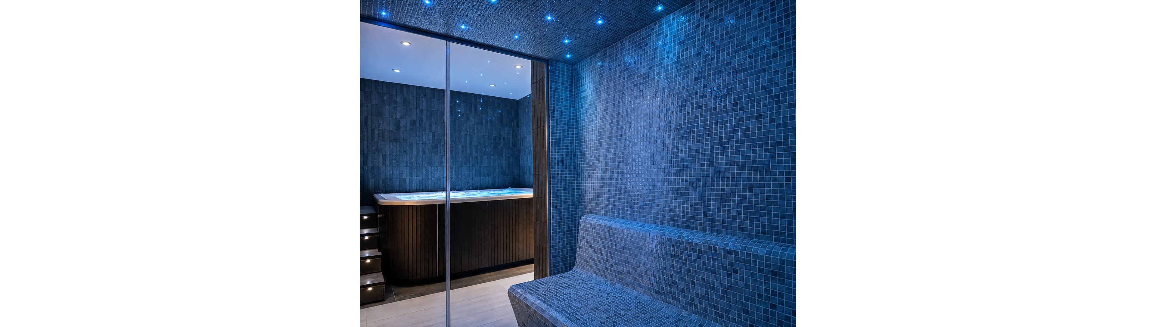 A contemporary spa interior features a blue tiled steam room with a built-in bench. Beyond a glass partition, a hot tub or jacuzzi is visible, framed by dark wood paneling and softly lit steps. Small star-like ceiling lights create a calming ambiance and reflect off the mosaic tiles. The overall scene suggests a luxurious wellness or relaxation space.