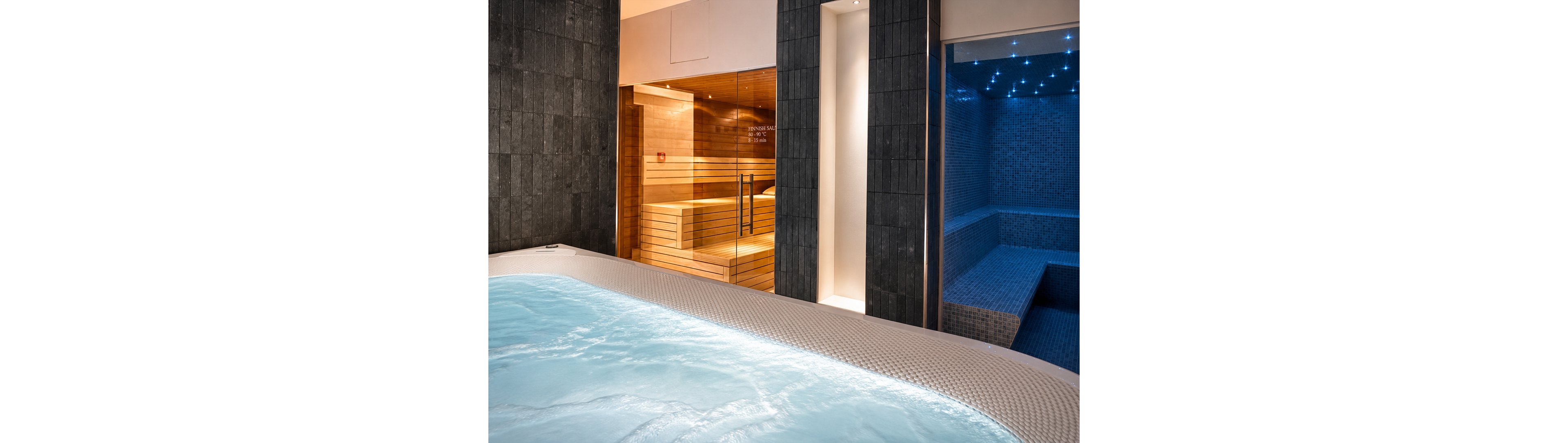 A modern spa interior features a bubbling jacuzzi pool in the foreground. Behind the pool, a warm wooden sauna room is visible through glass doors. To the right, a blue-tiled steam room glows with soft star-like ceiling lights. The scene highlights contrasting warm and cool tones, creating a relaxing wellness atmosphere.