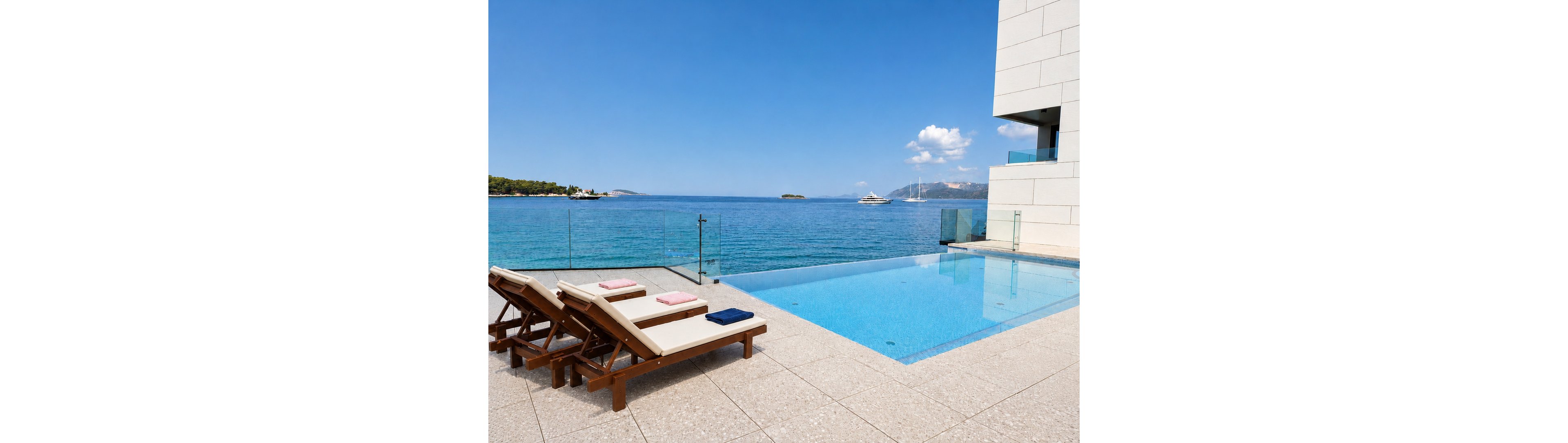 A modern infinity pool sits on a stone terrace overlooking a calm blue sea. Wooden lounge chairs with cushions and folded towels line the poolside, facing the ocean view. A glass railing separates the pool area from the water, while a contemporary white building frames the right side of the scene. In the distance, small islands, a yacht, and a partly cloudy sky enhance the serene coastal atmosphere.