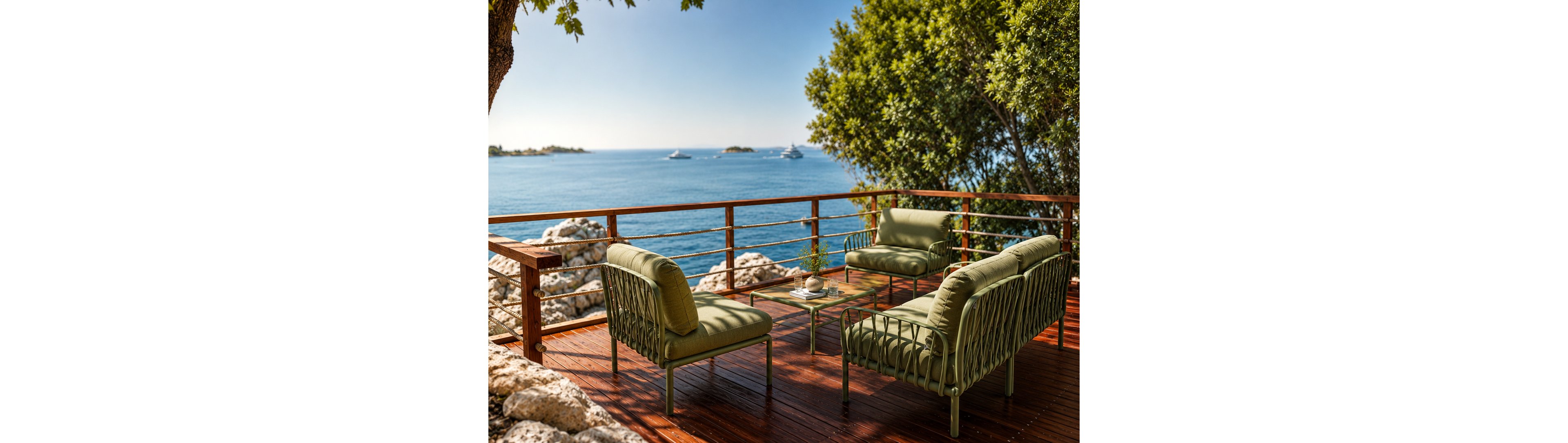 An outdoor terrace with modern green lounge chairs overlooks a calm blue ocean. The wooden deck is bordered by a simple metal railing and surrounded by rocky shoreline and lush trees. A low glass coffee table with small decorative items sits at the center of the seating area. In the distance, several boats and small islands are visible under clear, sunny skies.