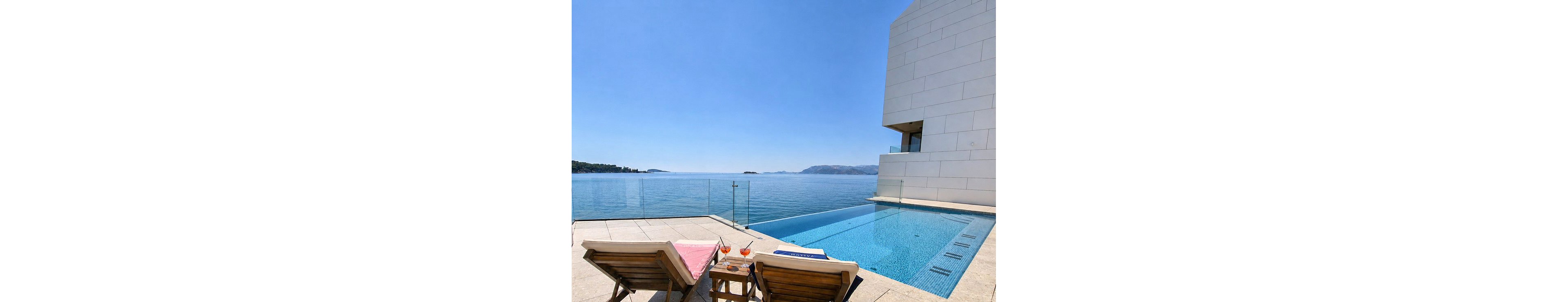 A contemporary waterfront terrace features an infinity pool overlooking a calm blue sea. Two wooden lounge chairs with pink towels face the water beside a small table holding colorful drinks. A minimalist white building rises on the right, framed by clear glass railings along the edge of the terrace. Distant islands and hazy mountains are visible under a bright, cloudless sky, creating a serene vacation atmosphere.