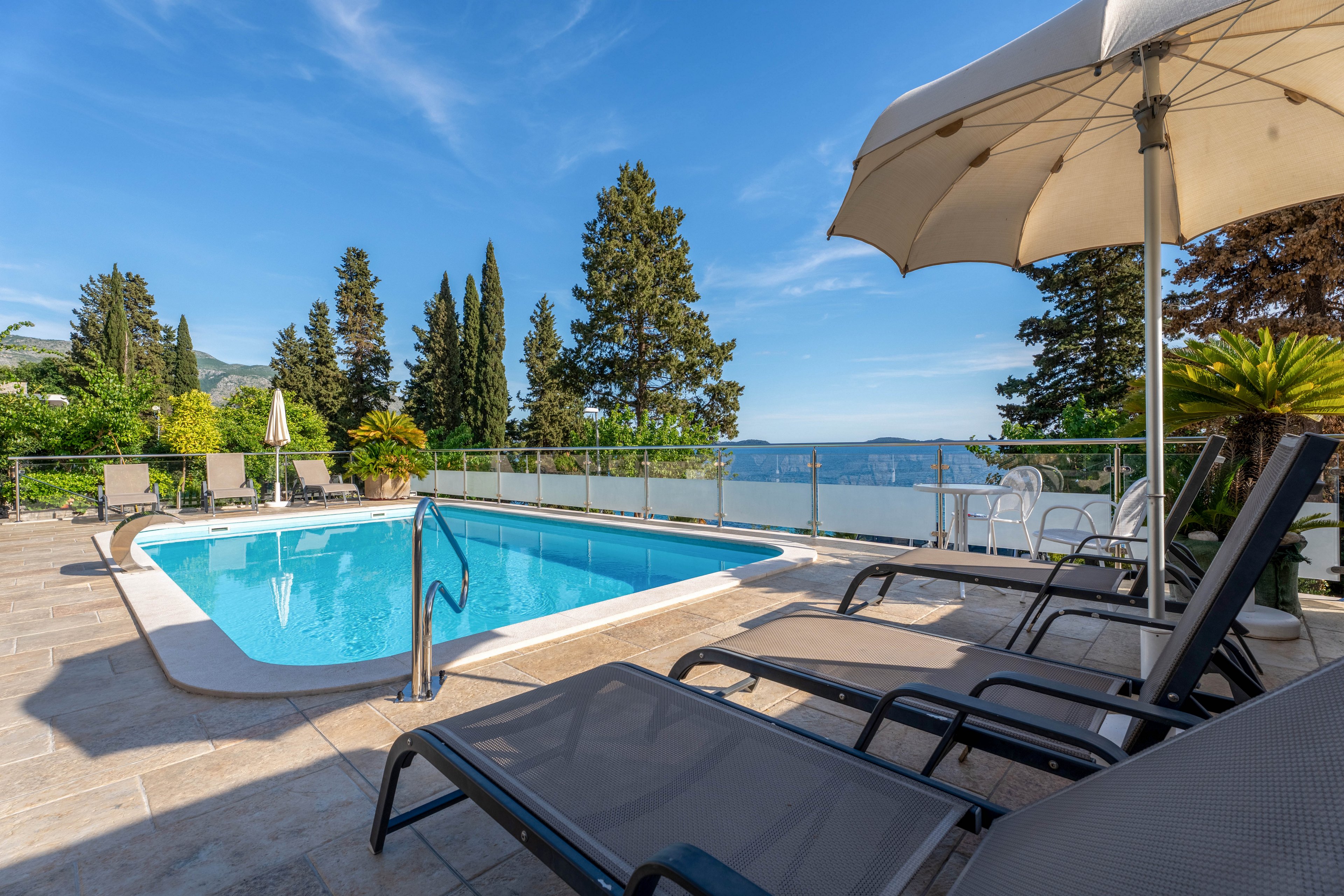 Outdoor swimming pool on a tiled terrace with sun loungers and a large umbrella in the foreground. Glass railing surrounds the pool area, opening to a scenic view of the sea and distant hills. Tall evergreen trees and lush greenery frame the setting under a clear blue sky. The scene suggests a relaxing vacation atmosphere at a seaside property.