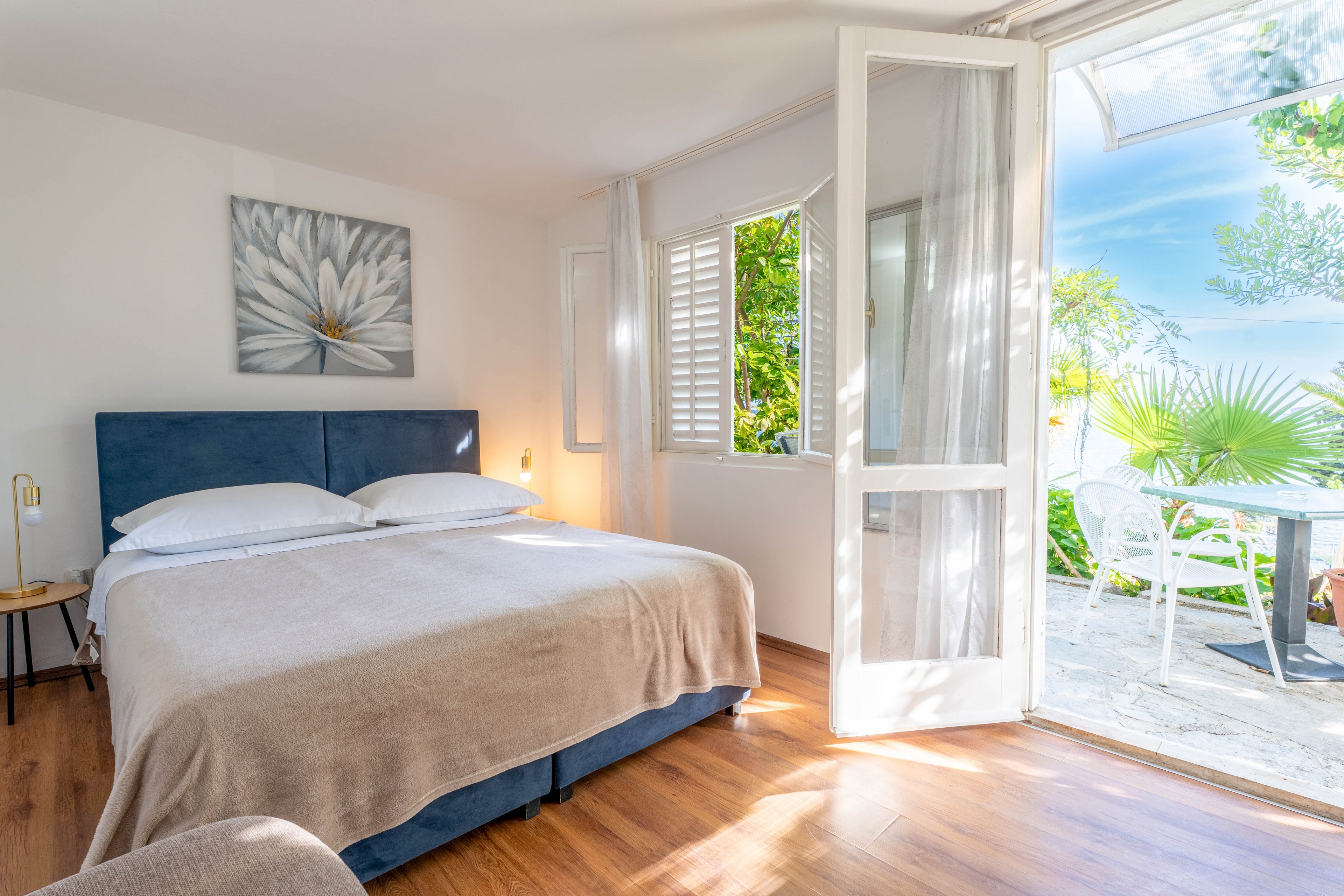 A sunlit bedroom features a double bed with white linens and a beige blanket on warm wooden flooring. A large flower artwork hangs above the bed, adding a soft decorative touch to the minimalist interior. Open windows and French doors reveal a lush garden terrace with outdoor seating and vibrant greenery. Natural light floods the room, creating an airy, relaxing atmosphere ideal for a vacation stay.