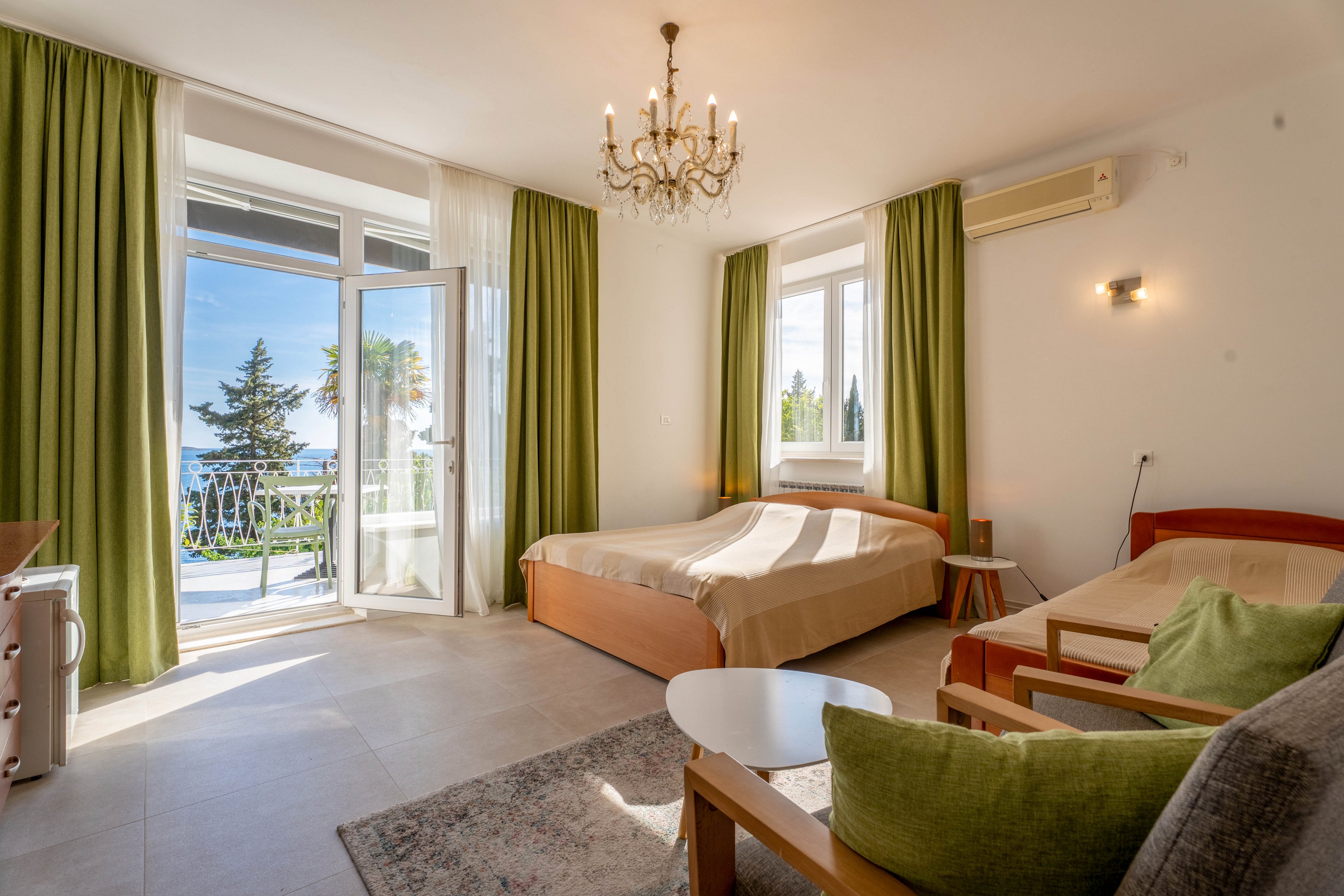 A sunlit hotel room features a double bed and a single bed arranged near large windows. Green curtains frame the open balcony door, revealing an outdoor view with trees and distant water. A small seating area with a sofa, cushions, and a round table sits on a rug in the foreground. Warm lighting from a chandelier and wall sconce enhances the airy, relaxed atmosphere.