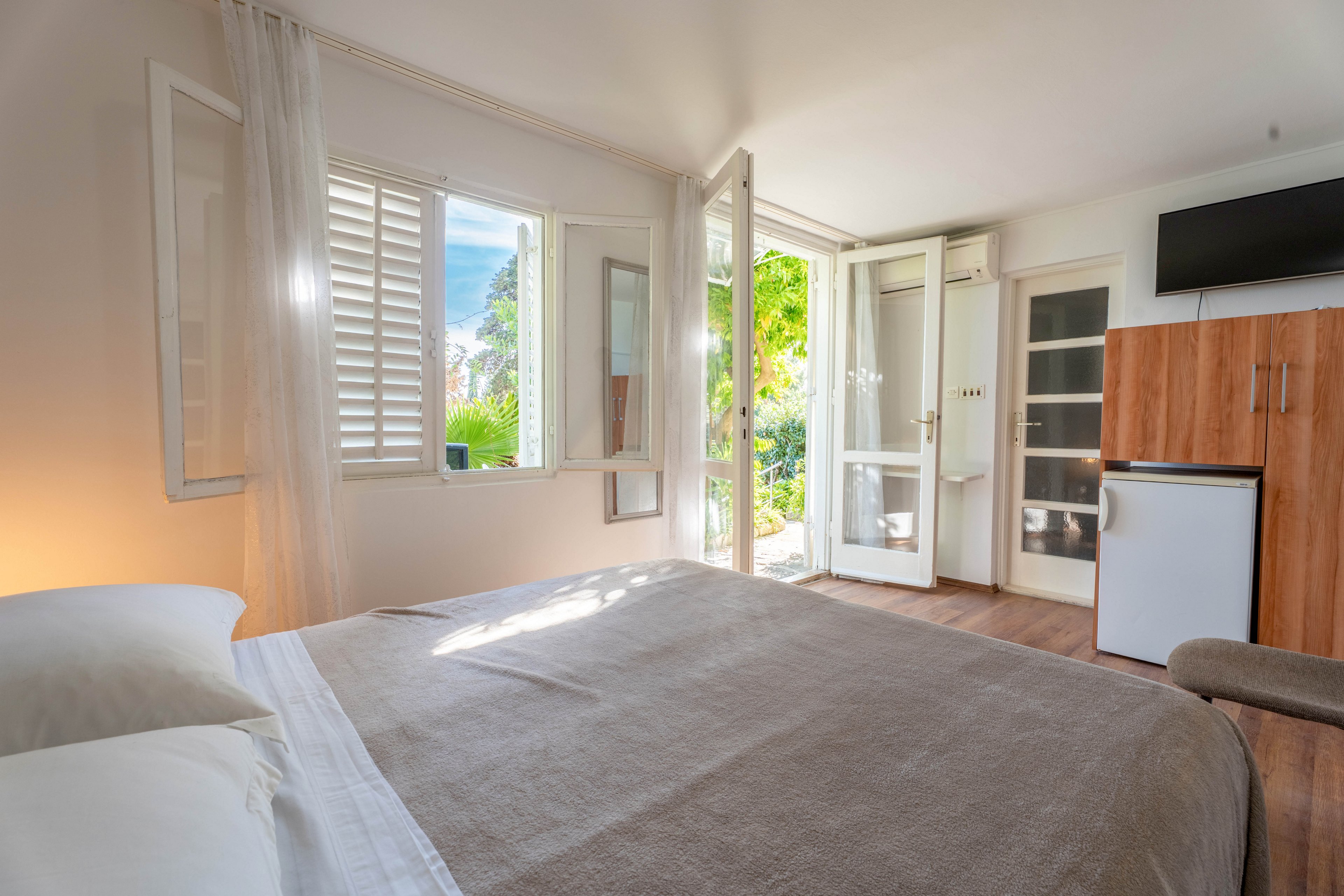Cozy guest bedroom with a neatly made double bed faces large windows and an open glass door. Sunlight streams in, illuminating light-colored walls, sheer curtains, and wooden flooring. A small white refrigerator and wooden storage cabinet stand against the wall beneath a mounted television. Outside greenery is visible through the open door and shutters, creating a fresh, inviting atmosphere.