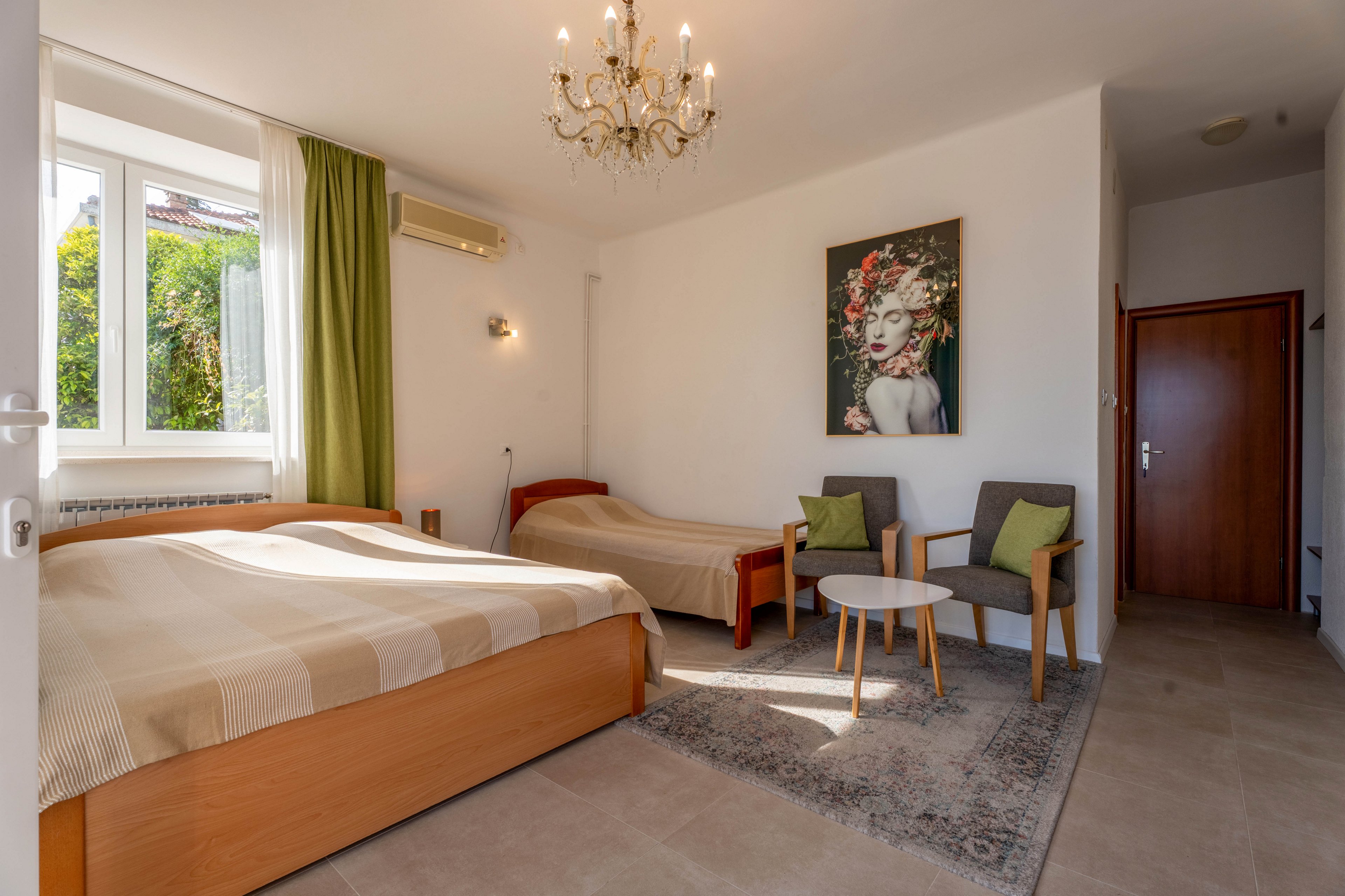 A sunlit guest bedroom features a large double bed and a smaller single bed with neutral bedding. Two modern armchairs with green cushions and a small round table sit on a patterned rug, creating a cozy seating area. A decorative chandelier hangs from the ceiling, and a large window with green curtains lets in natural light and garden views. A colorful portrait artwork on the wall adds character to the simple, clean interior.