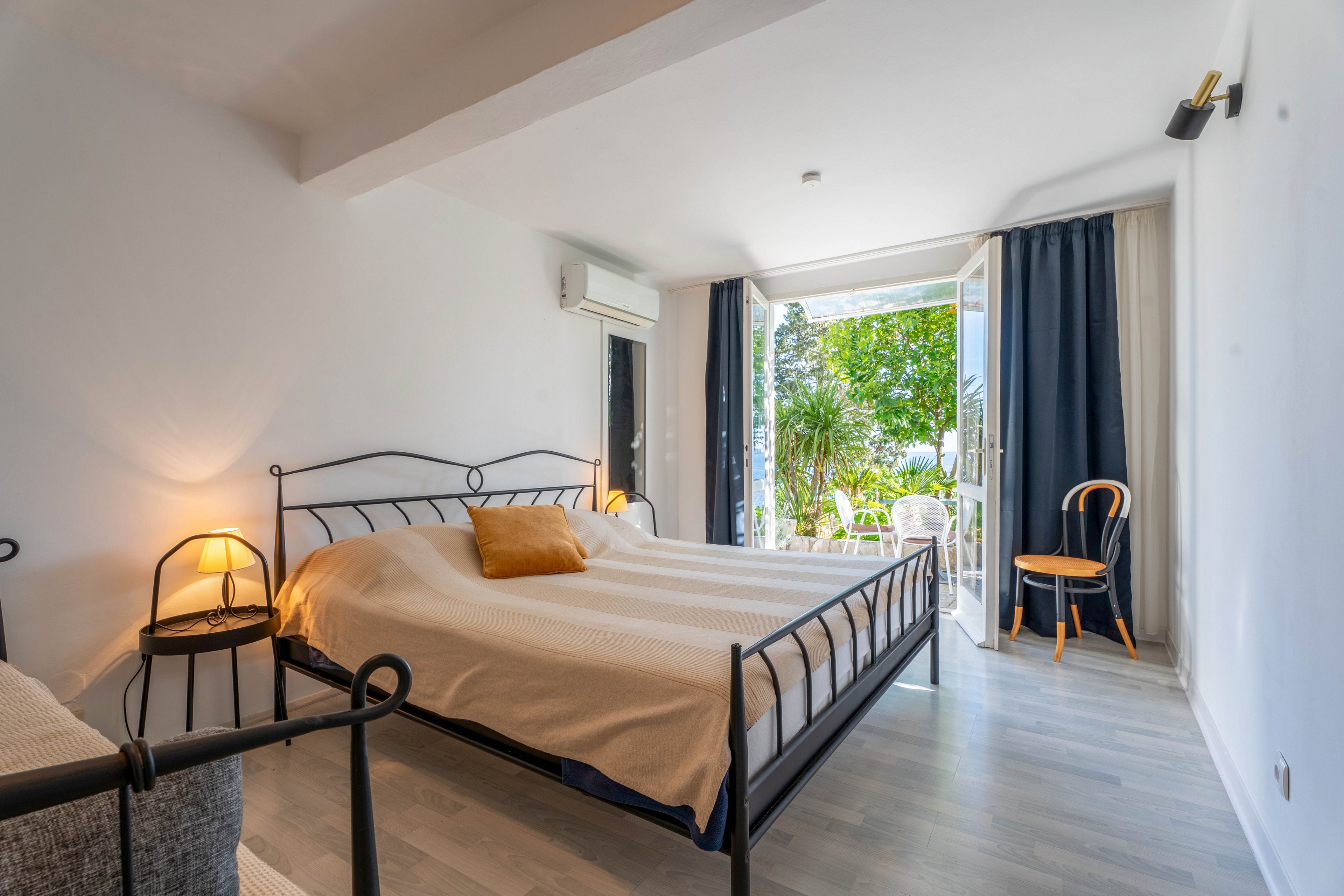 A contemporary bedroom features a double bed with a metal frame and light striped bedding accented by a mustard-colored pillow. Warm bedside lamps create a cozy atmosphere against the clean white walls. Large open glass doors reveal a lush green garden and outdoor seating area, bringing natural light into the room. A wooden chair, air conditioning unit, and dark curtains complete the simple, modern decor.