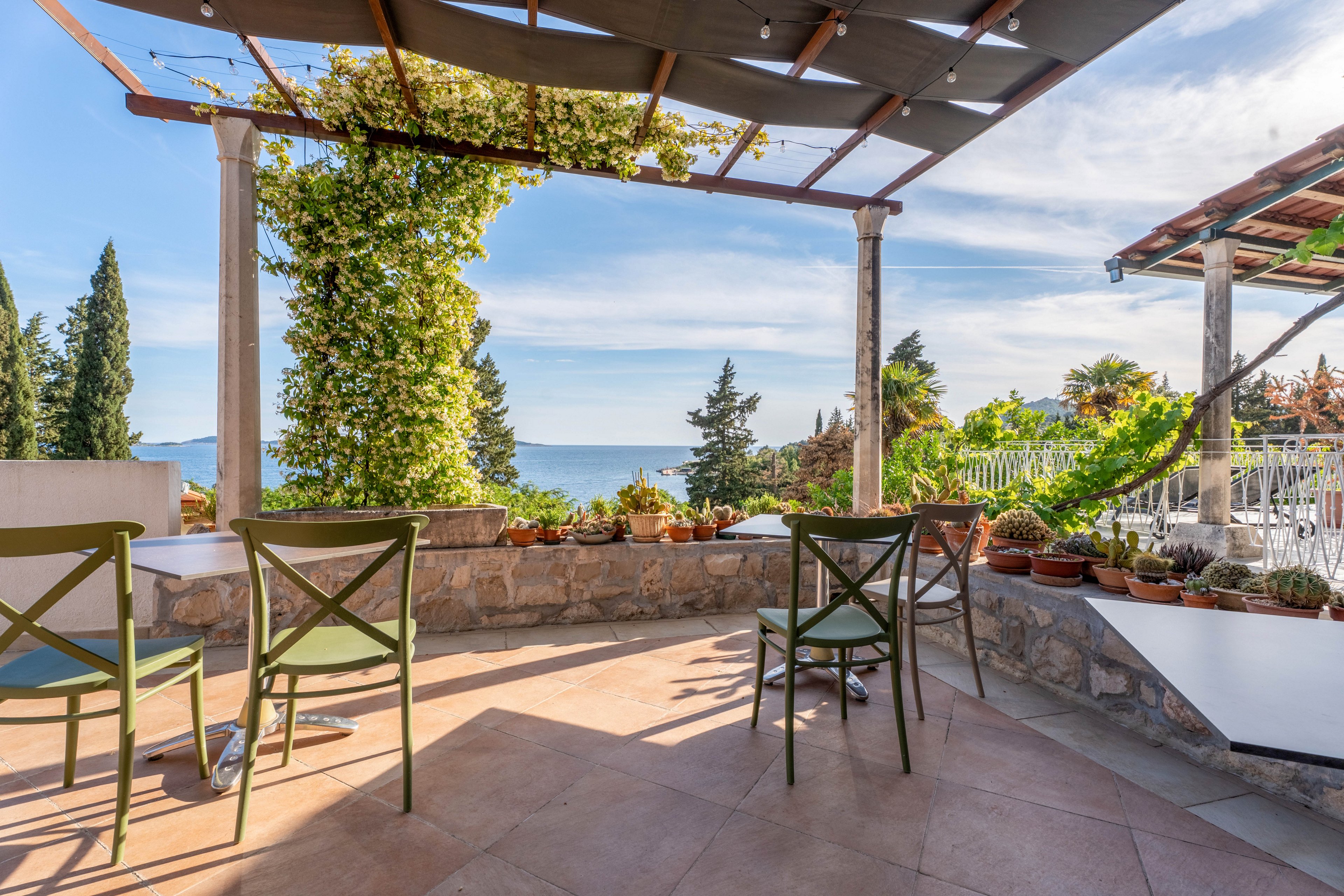 An open terrace features several green chairs and a stone wall overlooking a distant sea view. Potted plants line the edge of the patio, and a pergola with climbing vines provides partial shade. Tall trees and lush vegetation surround the area, creating a tranquil, Mediterranean-style setting. The bright daylight and clear sky give the scene a warm, inviting atmosphere.