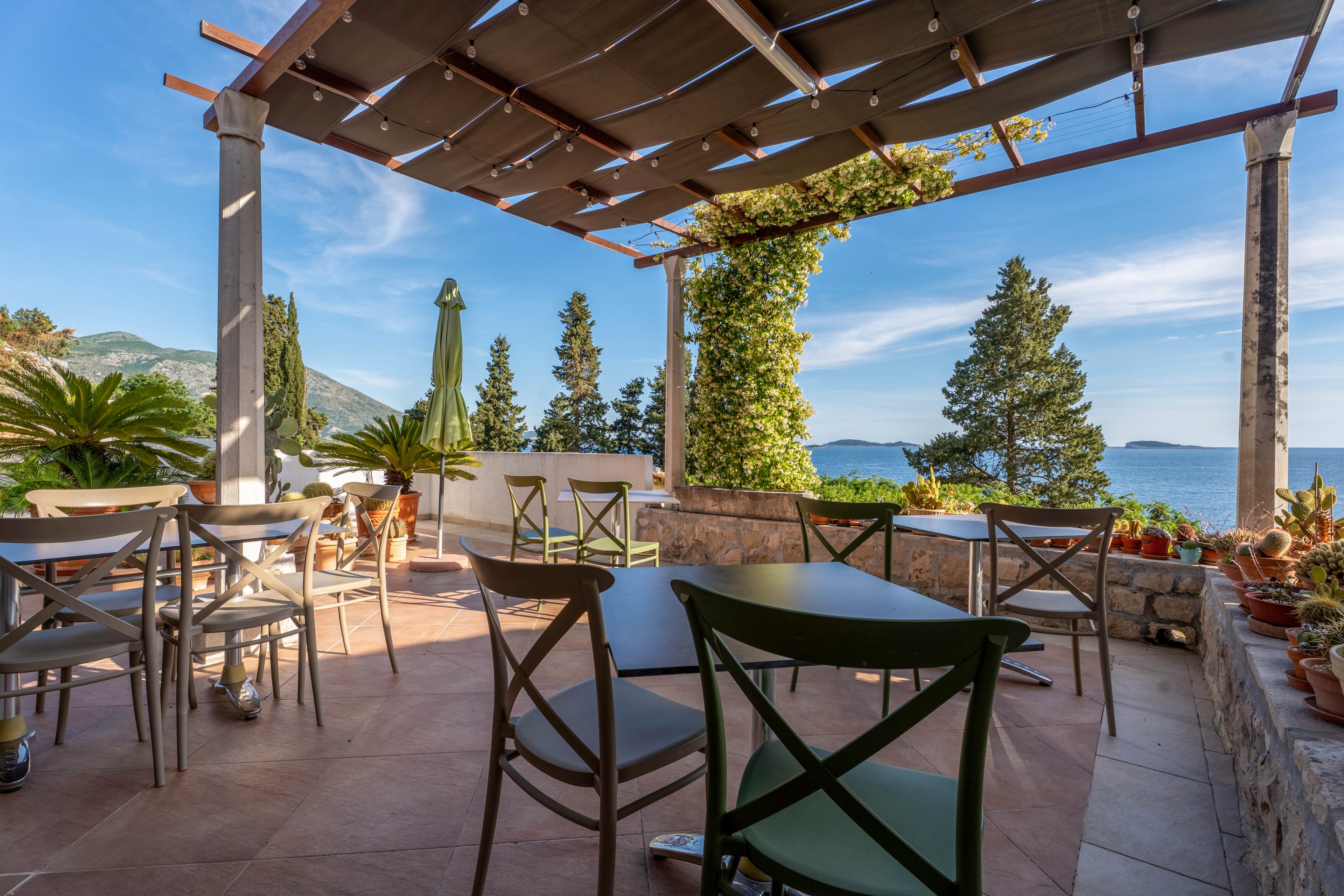 An open-air terrace café features empty tables and chairs under a partial pergola roof. The setting overlooks a scenic coastline with blue sea, distant islands, and green hills. Potted plants and climbing vines add natural accents around the stone walls and columns. Bright daylight and clear skies create a relaxed, inviting atmosphere for outdoor dining.