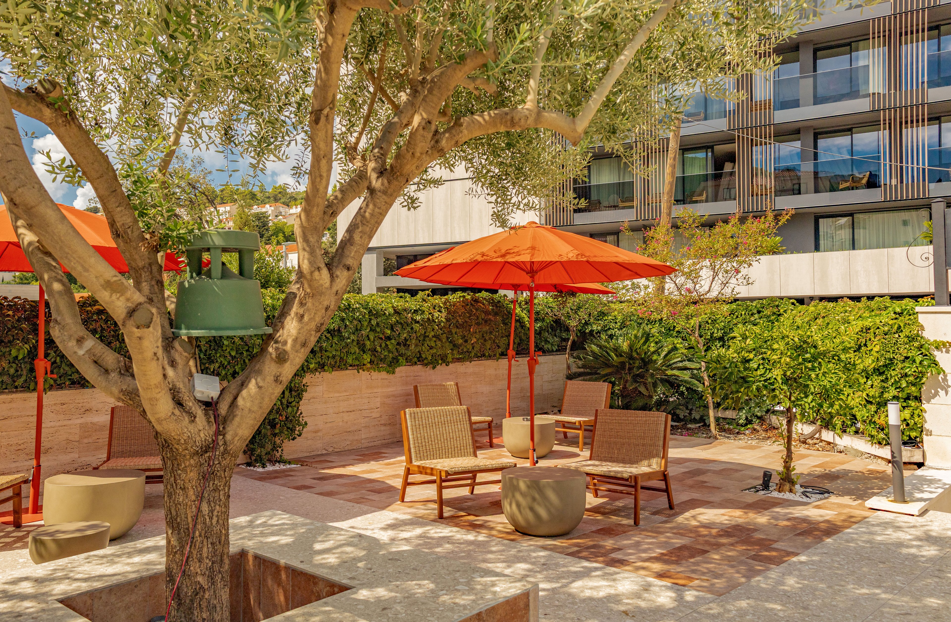 A contemporary outdoor courtyard features lounge seating beneath bright orange umbrellas. Wooden chairs and small tables are arranged on a sunlit patio surrounded by lush green hedges and trees. A modern multi-story building with balconies forms the backdrop. The scene suggests a relaxed, resort-like atmosphere ideal for socializing or resting outdoors.