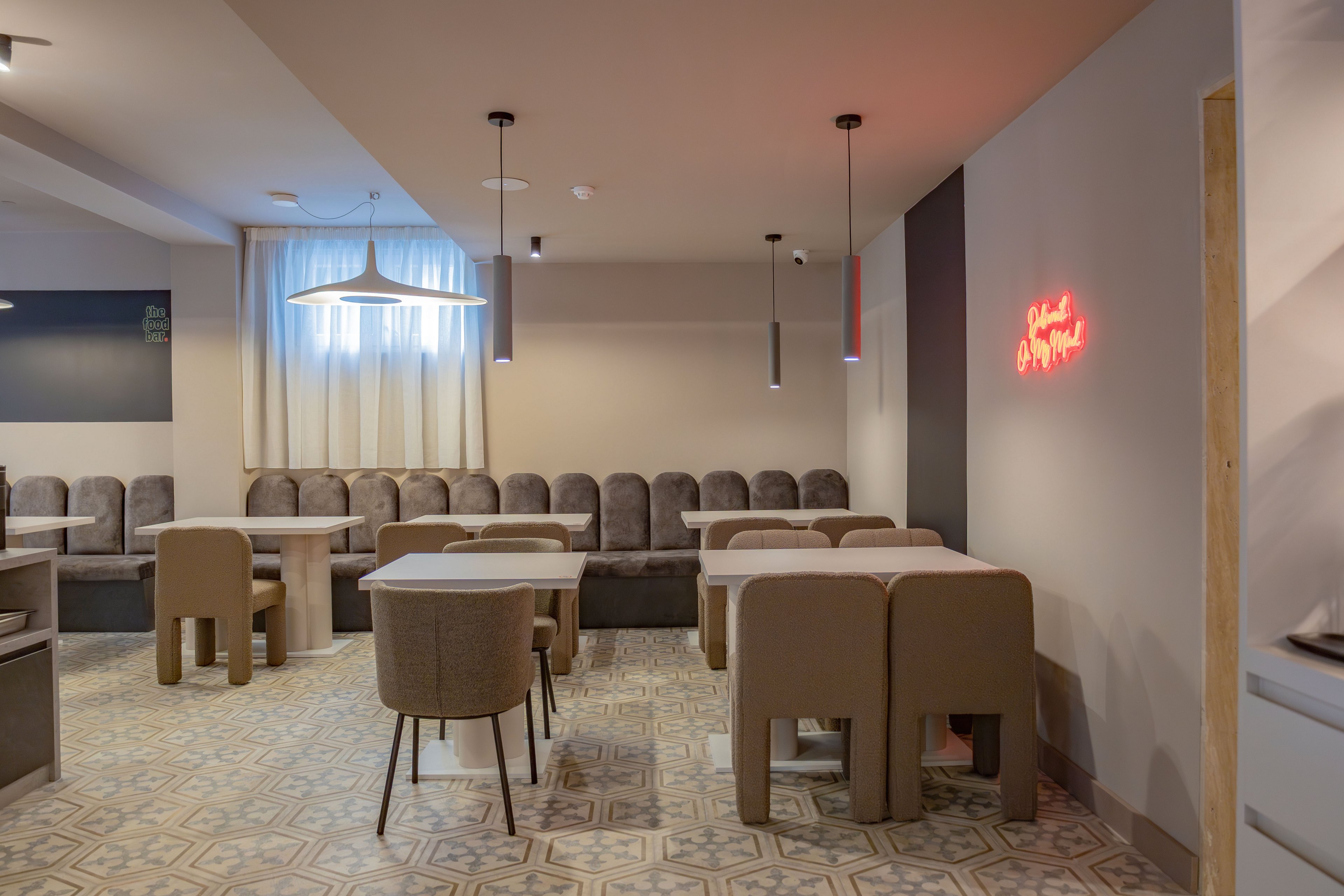 A contemporary cafe interior features rows of cushioned banquette seating and simple rectangular tables. Neutral beige and gray tones dominate the space, complemented by patterned floor tiles. Pendant lights hang from the ceiling, including a large sculptural fixture and several slim cylindrical lamps. A red neon sign glows on the wall, adding a warm accent to the otherwise minimalist decor.