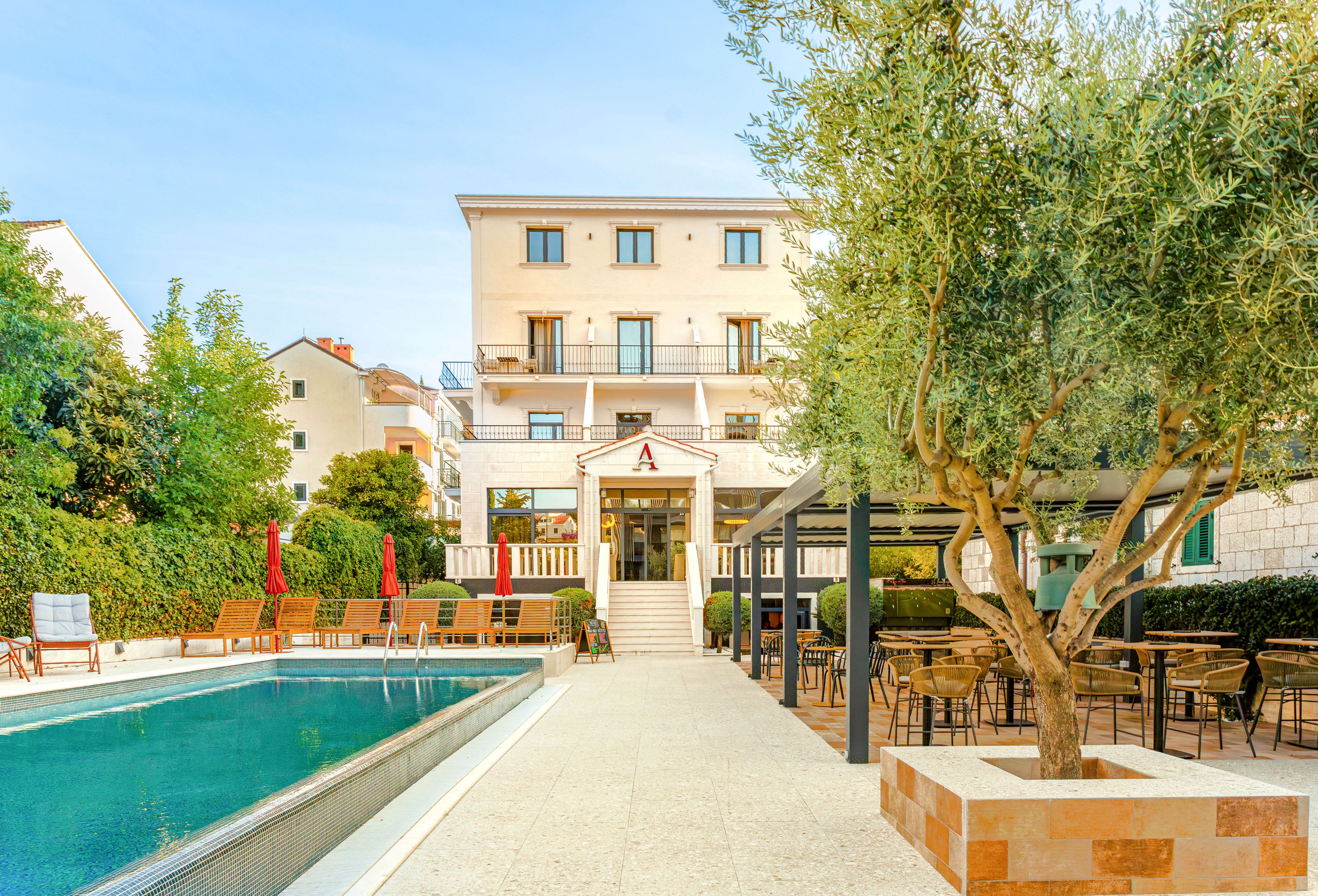 A bright hotel courtyard features a long rectangular swimming pool surrounded by lounge chairs and umbrellas. The multi-story building in the background has balconies overlooking the pool area. On the right side, an outdoor seating area with tables and chairs sits beneath a pergola and mature trees. The scene is captured in warm daylight, creating a relaxed vacation atmosphere.