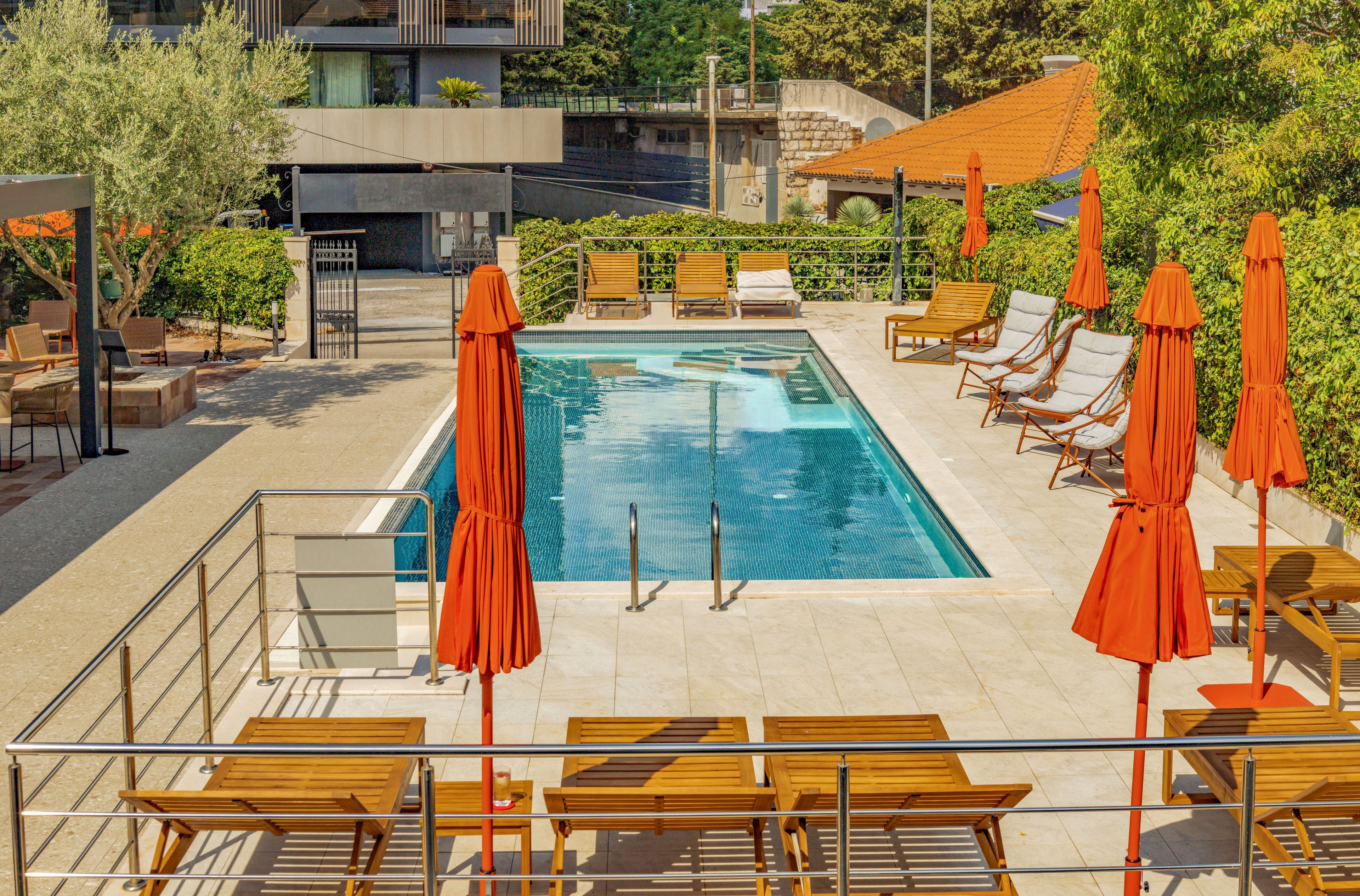 A rectangular outdoor swimming pool is surrounded by wooden sun loungers and bright orange umbrellas. The pool area is set within a modern hotel or apartment complex, with greenery and hedges enclosing the space. Metal railings and a tiled deck frame the pool, creating a clean and organized layout. Warm sunlight and vivid colors give the scene a relaxed, vacation-like atmosphere.