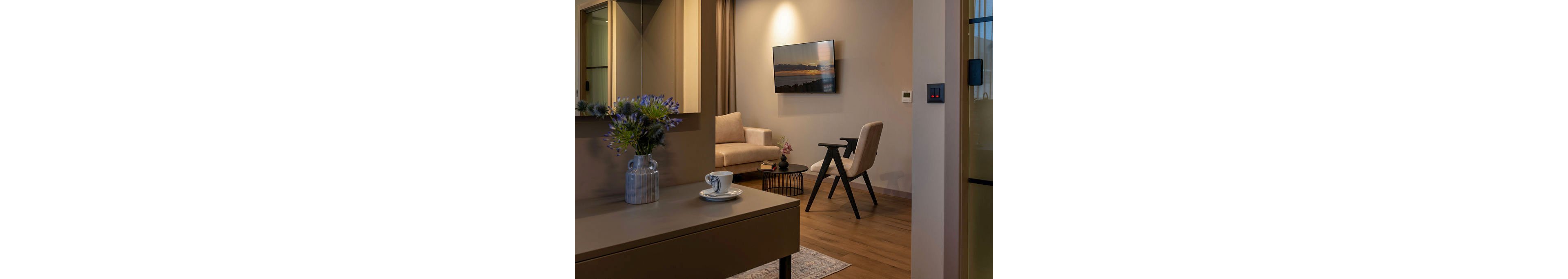 A contemporary living room features a beige armchair and a matching sofa arranged around a small round side table. A wall-mounted television displays a scenic landscape under a focused spotlight, adding visual interest to the neutral walls. In the foreground, a sleek console table holds a vase of flowers and a cup with saucer, enhancing the welcoming atmosphere. Warm wooden flooring and soft lighting create a calm, comfortable interior setting.