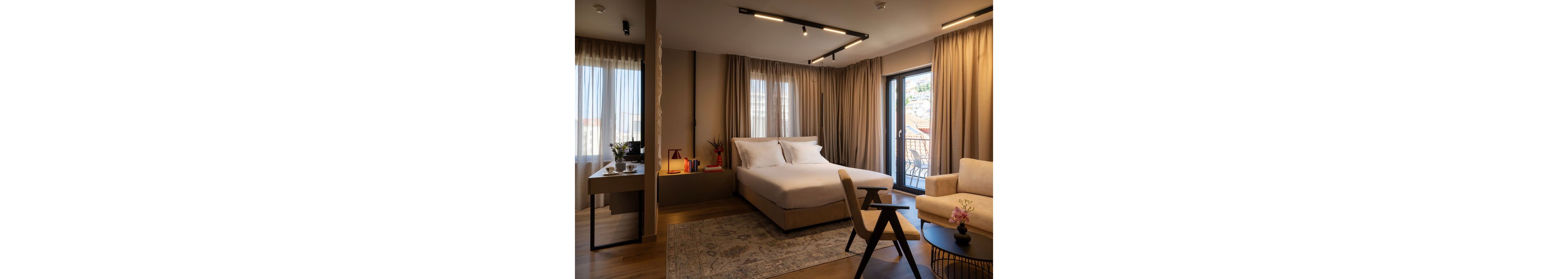 A contemporary hotel bedroom features a neatly made double bed with white linens and soft beige upholstery. Floor-to-ceiling curtains frame large windows and a glass door leading to a balcony with an urban view. Warm ambient lighting, a patterned area rug, and wooden flooring create a welcoming atmosphere. A small desk, armchair, side table with flowers, and decorative books add comfort and functional details to the space.