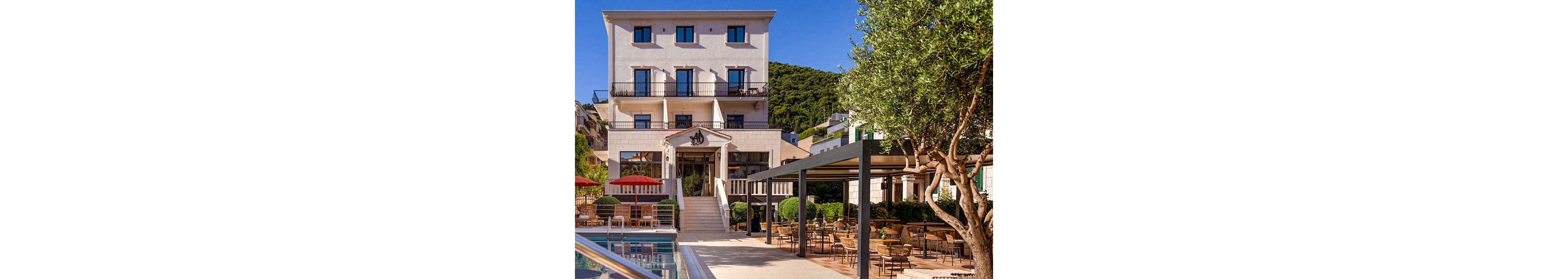 A multi-story hotel building overlooks a sunlit courtyard with an outdoor swimming pool in the foreground. Numerous tables and chairs are arranged on the terrace, suggesting an open-air dining or lounge area. Red umbrellas and manicured greenery add color and structure to the scene, while surrounding hills and trees frame the property. The warm daylight and clear sky create a relaxed, inviting vacation atmosphere.