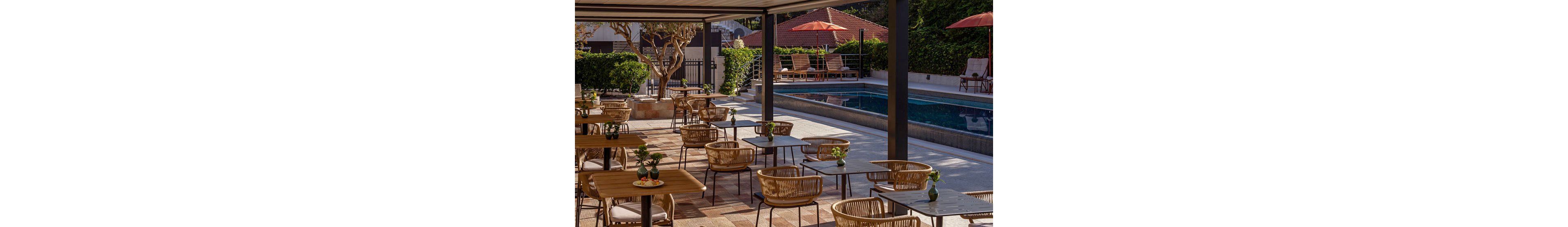 An open-air cafe terrace with wooden tables and woven chairs sits beside a long rectangular swimming pool. The scene is shaded by a pergola, while sun loungers and umbrellas line the poolside in the background. Potted plants and small floral arrangements on the tables add a touch of greenery to the warm, tiled flooring. The overall atmosphere is relaxed and inviting, suggesting a resort or boutique hotel setting.