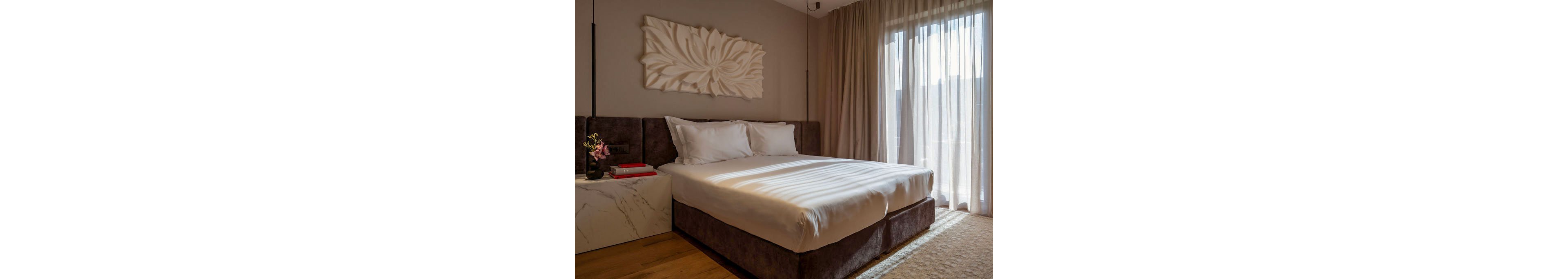A neatly made double bed with white linens sits in a modern bedroom interior. Soft daylight filters through sheer curtains covering a large window, creating a warm and inviting atmosphere. A marble-look bedside table holds a small flower arrangement and books, while a large sculptural wall art piece decorates the headboard area. The room features neutral tones, wooden flooring, and a cozy textured rug for a contemporary, comfortable feel.