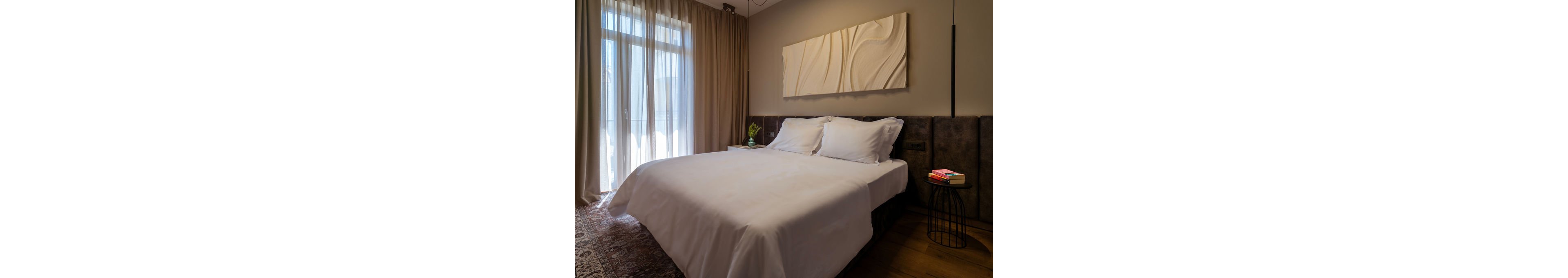 A neatly made double bed with white linens is the focal point of a modern bedroom. Soft natural light filters through sheer curtains covering a large window. A textured abstract artwork hangs above the bed, and a small round side table holds books and decor. Warm wood flooring and a patterned rug add to the cozy, contemporary atmosphere.