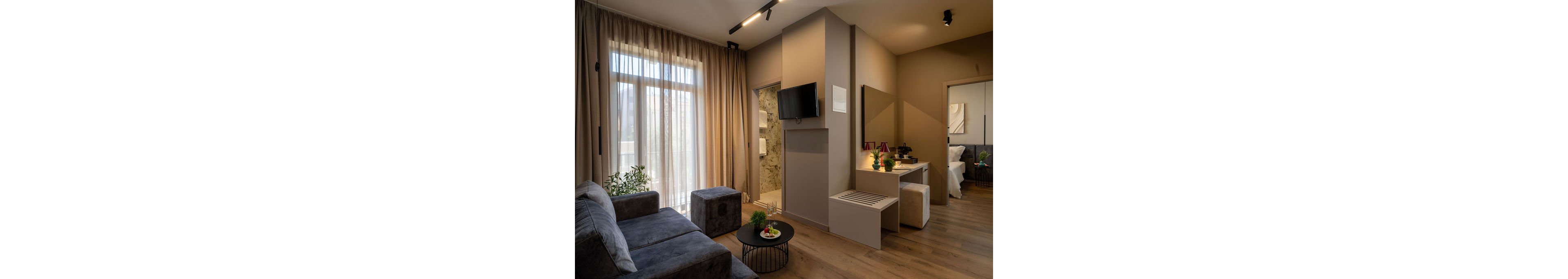 Contemporary hotel suite interior featuring a gray sofa and small round coffee table in the living area. Floor-to-ceiling curtains cover large windows, allowing soft natural light into the room. A wall-mounted television faces the seating area, with a vanity desk and mirror extending toward the bedroom. Warm neutral tones, wooden flooring, and subtle decorative accents create a comfortable, inviting atmosphere.
