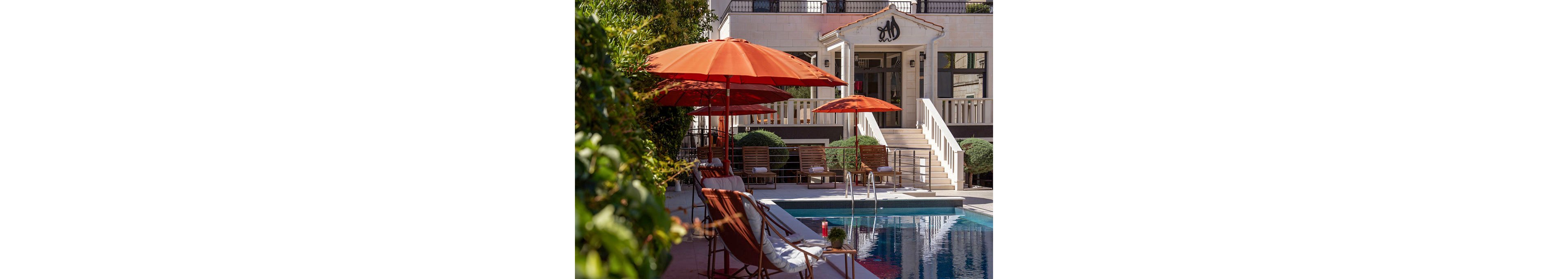 A stylish outdoor pool area is set in front of a light-colored building with a staircase and balcony. Bright orange umbrellas shade lounge chairs arranged along the poolside, creating a relaxed vacation atmosphere. Green shrubs and trees frame the scene, while a metal pool ladder leads into the clear blue water. A red beverage can and towels on the chairs add small pops of color and detail to the composition.