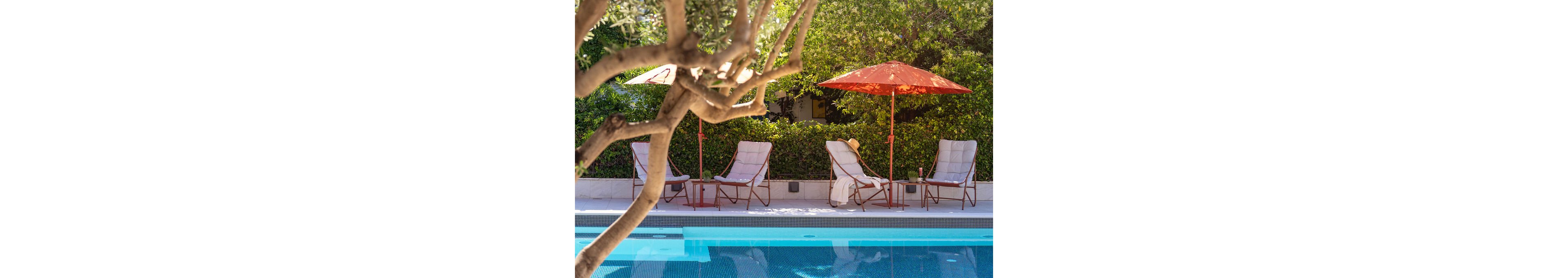 A tranquil outdoor poolside area features several sling lounge chairs arranged along a tiled deck. Bright orange umbrellas provide shade, with small side tables placed between the chairs. A sun hat rests on one of the chairs, suggesting a relaxed vacation atmosphere. Lush green hedges and trees surround the space, creating a private, inviting retreat.