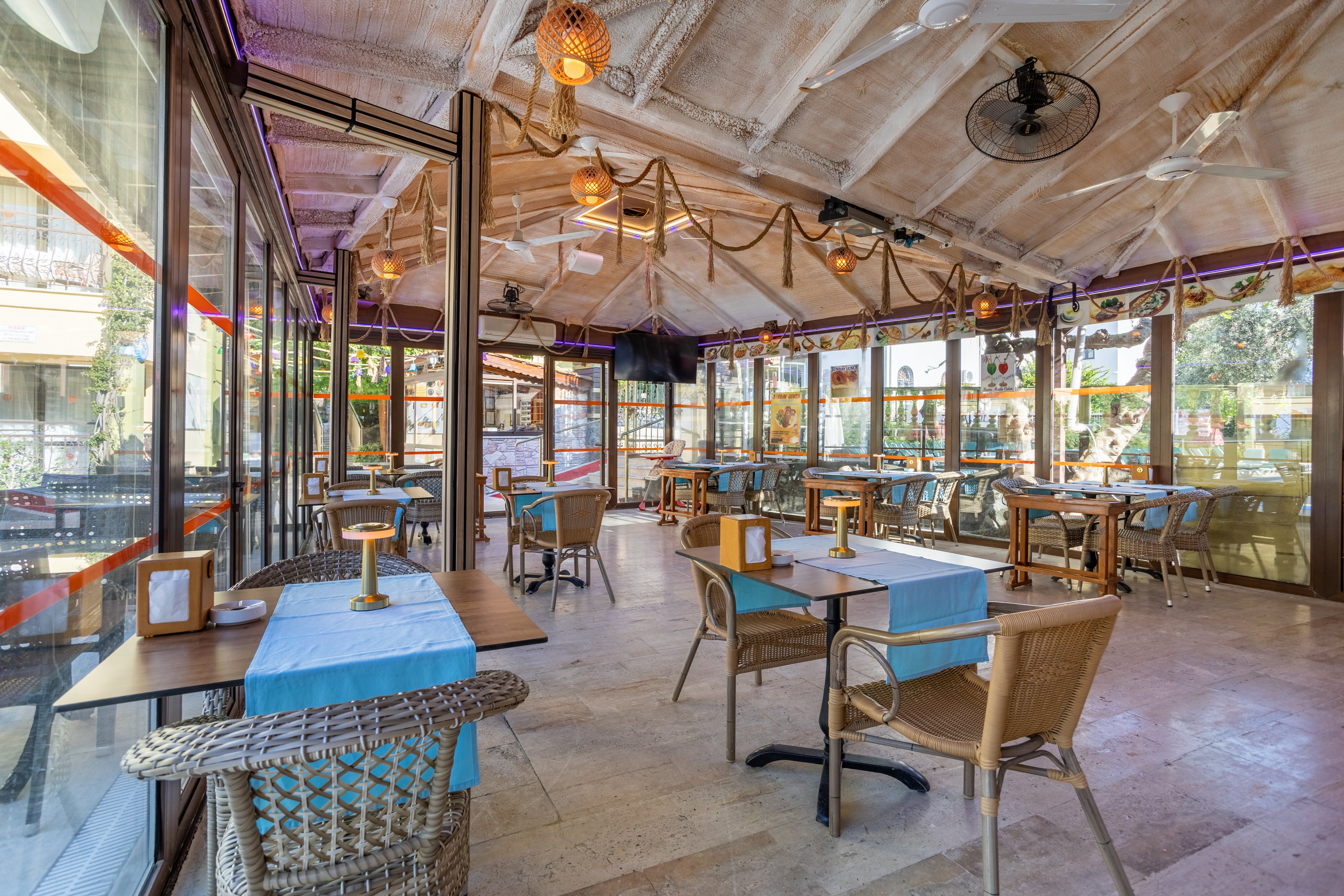 Spacious restaurant seating area with wicker chairs and small square tables covered in light blue tablecloths. The room features large glass windows on both sides, allowing abundant natural light and views of greenery outside. Warm hanging lights, ceiling fans, and decorative ropes create a cozy coastal ambiance. The space is empty of people, emphasizing the clean, airy, and inviting atmosphere.