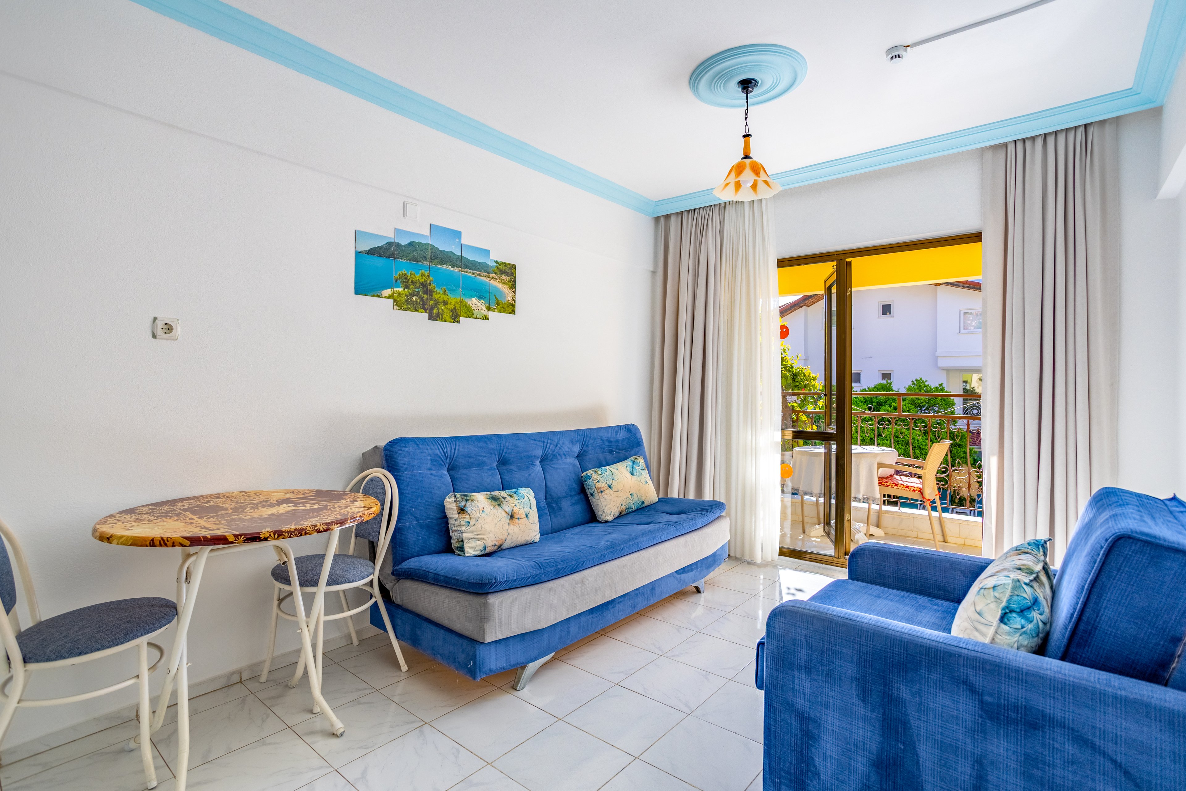 Cozy vacation apartment living room featuring blue sofas and patterned cushions. A small round dining table with two chairs sits beside the seating area. Large sliding glass doors open to a sunny balcony with outdoor chairs and greenery. Light tile flooring, white walls, and soft curtains create an airy, relaxed atmosphere.
