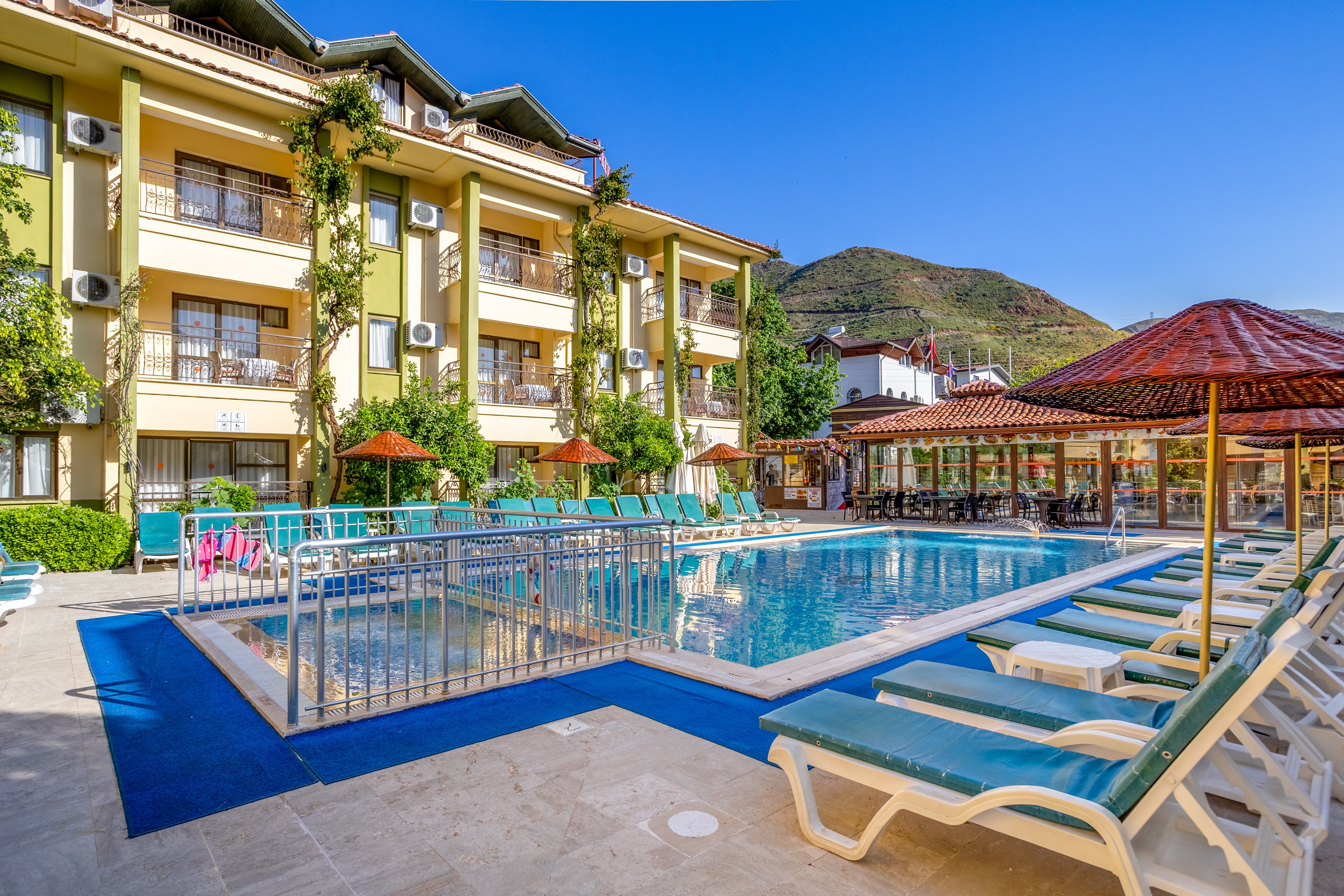 An inviting outdoor swimming pool sits at the center of a sunny hotel courtyard. Lounge chairs and umbrellas surround the pool, offering spaces for guests to relax. The multi-story building features balconies with greenery and exterior air-conditioning units. In the background, tree-covered hills rise under a clear blue sky, enhancing the resort-like atmosphere.