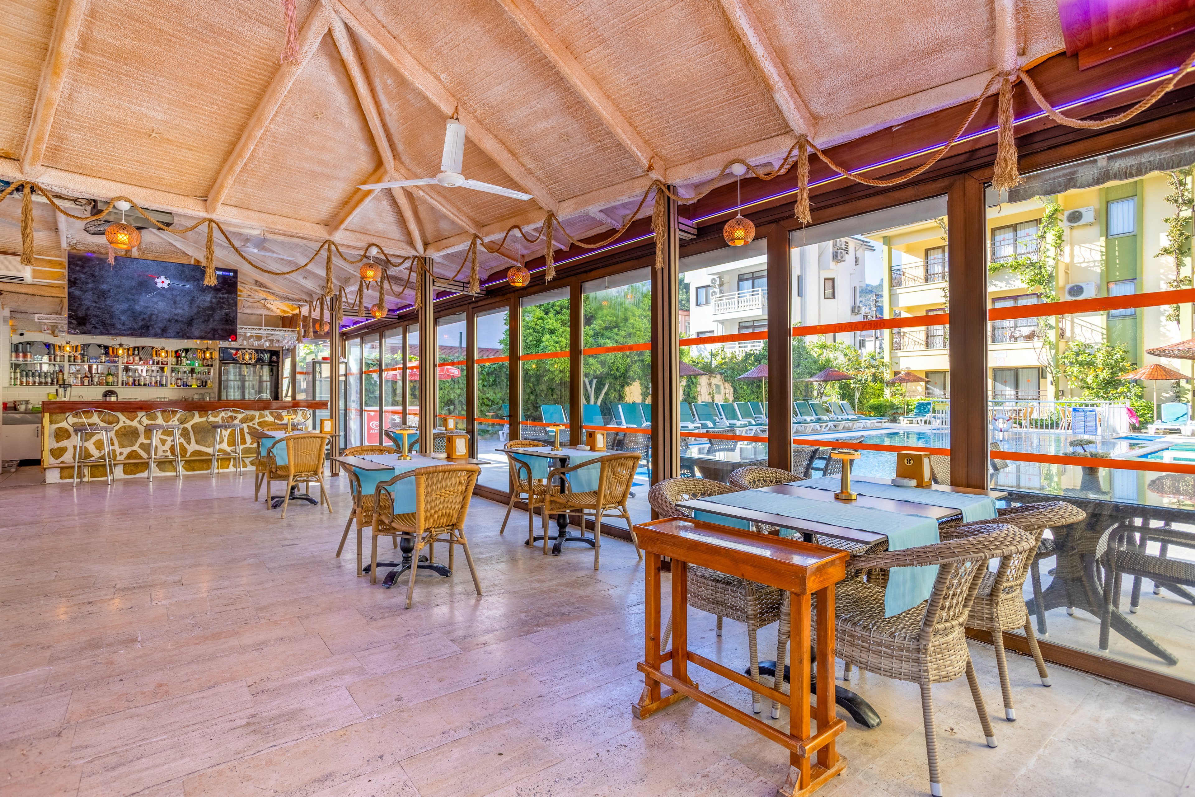 An open-air restaurant features neatly arranged tables and wicker chairs beside large glass windows. The interior has a high wooden ceiling with hanging lights and nautical rope decor. Through the windows, a sunny outdoor pool area and surrounding buildings are visible. The space appears calm and ready for guests, suggesting a relaxed vacation or resort atmosphere.