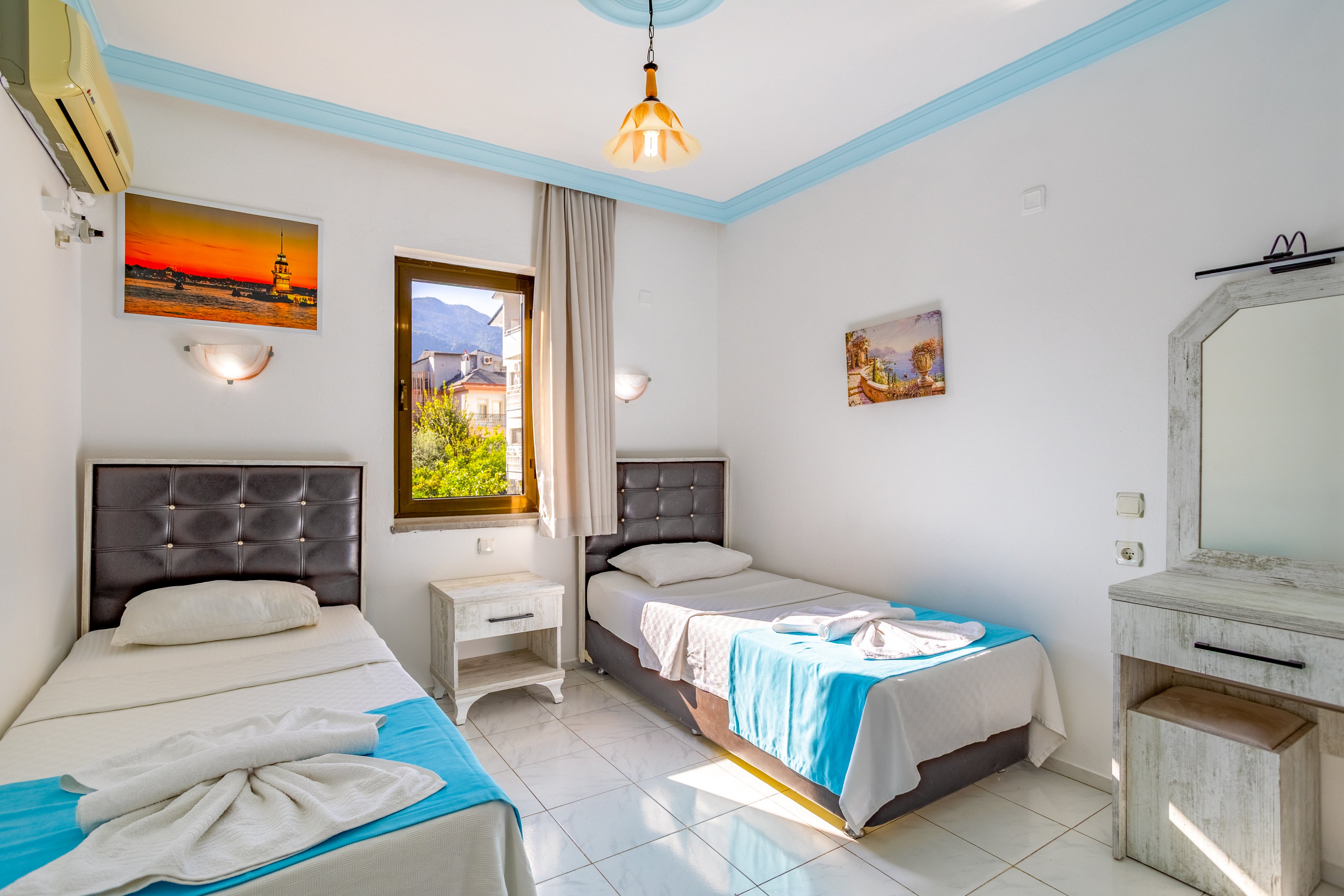 A clean, sunlit bedroom features two single beds with tufted gray headboards and white linens accented by light blue throws. The room has white tiled floors, a small bedside table, and a simple dressing table with a mirror. Natural light streams through the central window, revealing a scenic view of mountains and nearby buildings. Wall art, soft lighting fixtures, and pastel blue trim create a fresh, inviting atmosphere suitable for a hotel or guesthouse stay.