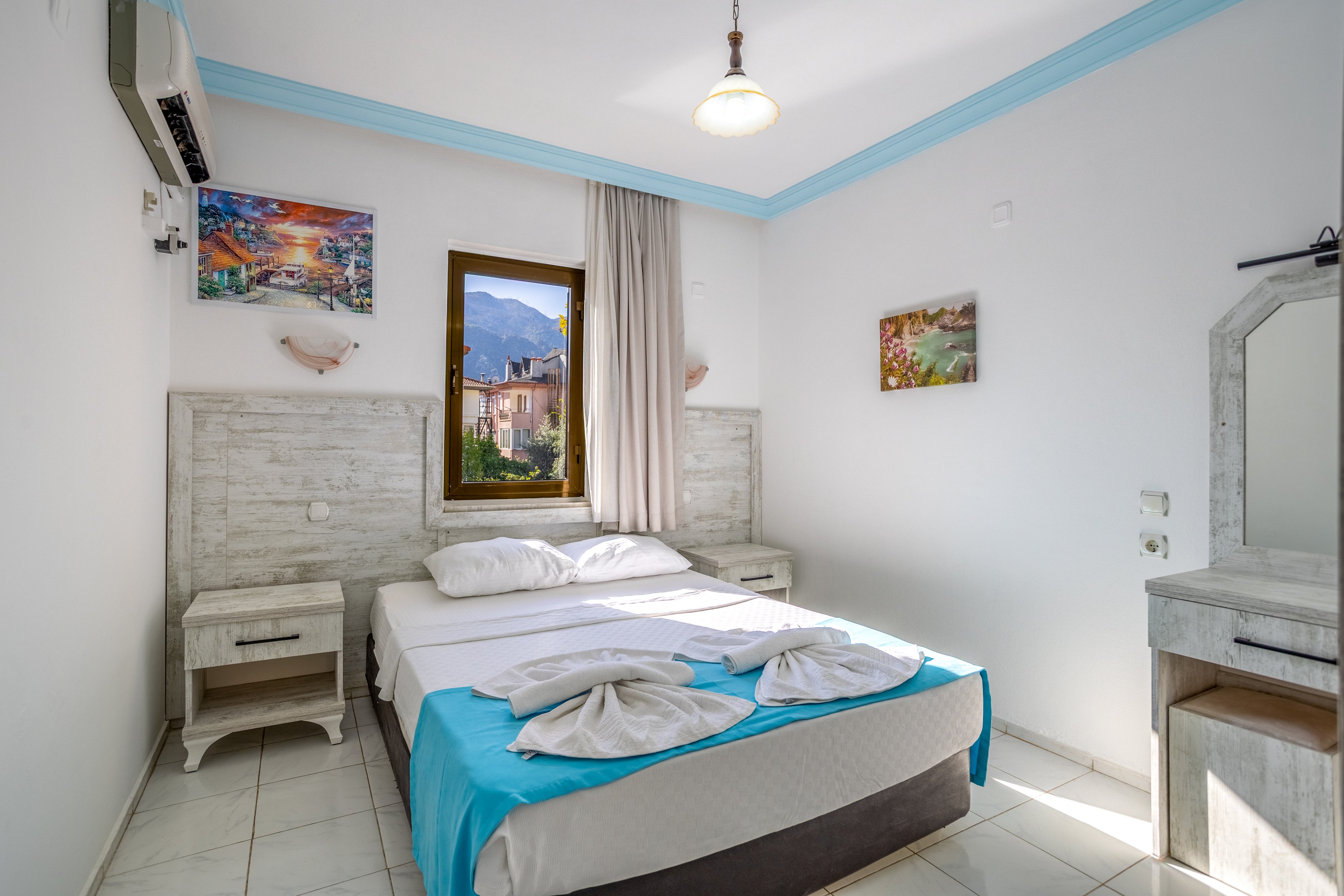 A neatly made double bed with white linens and a turquoise throw stands in the center of a bright hotel bedroom. The room features light wooden furniture, including bedside tables and a dressing table with mirror. A window between two headboards reveals a scenic view of mountains and buildings outside. Colorful wall art, tiled flooring, and a ceiling light complete the clean, minimalist interior.