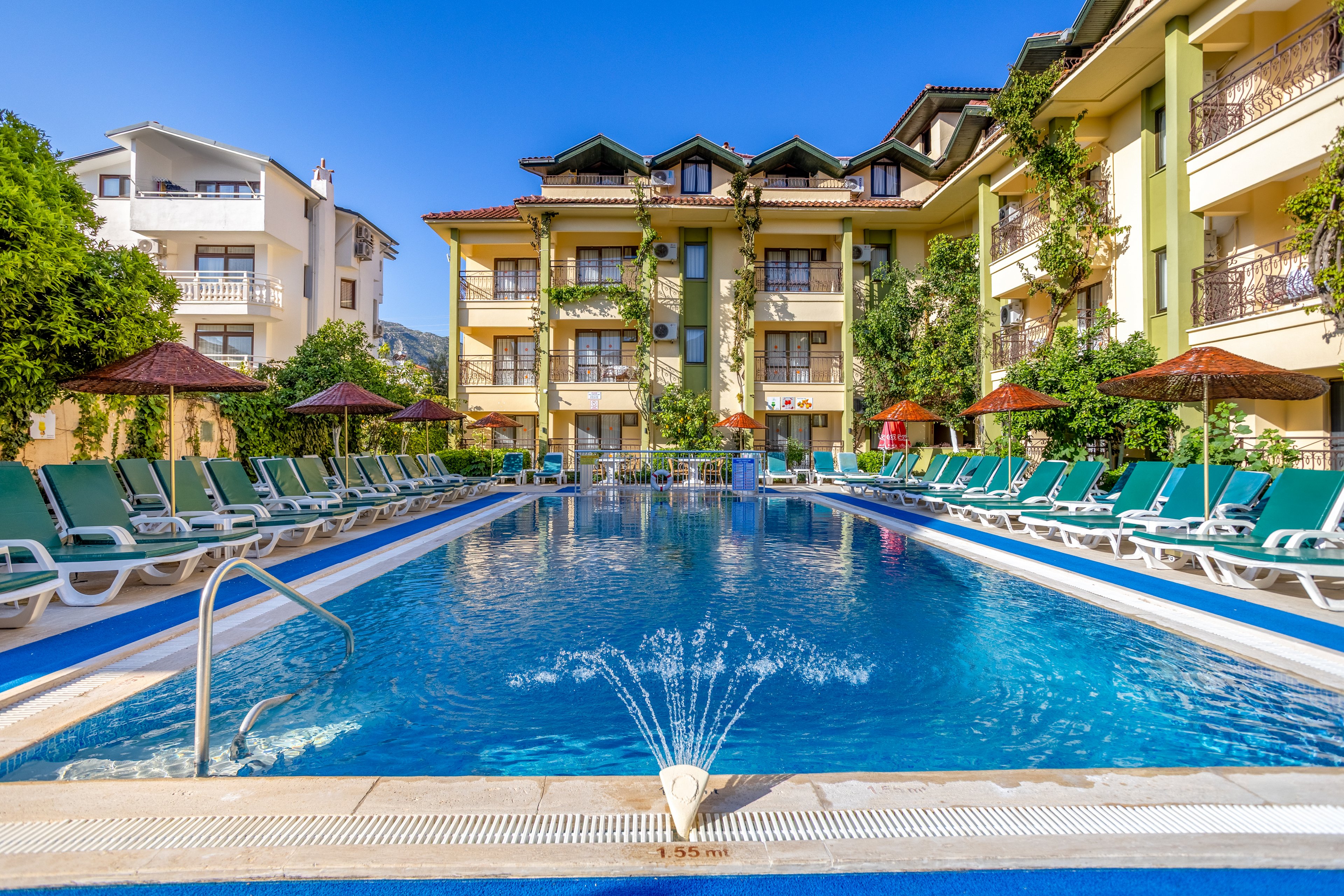 An outdoor hotel swimming pool is surrounded by rows of green sun loungers and parasols. The clear blue water features a small fountain spray at the shallow end, with metal handrails for easy access. Multi-story resort buildings with balconies and lush greenery frame the pool area under a bright blue sky. The scene suggests a relaxing vacation atmosphere in a warm, sunny climate.