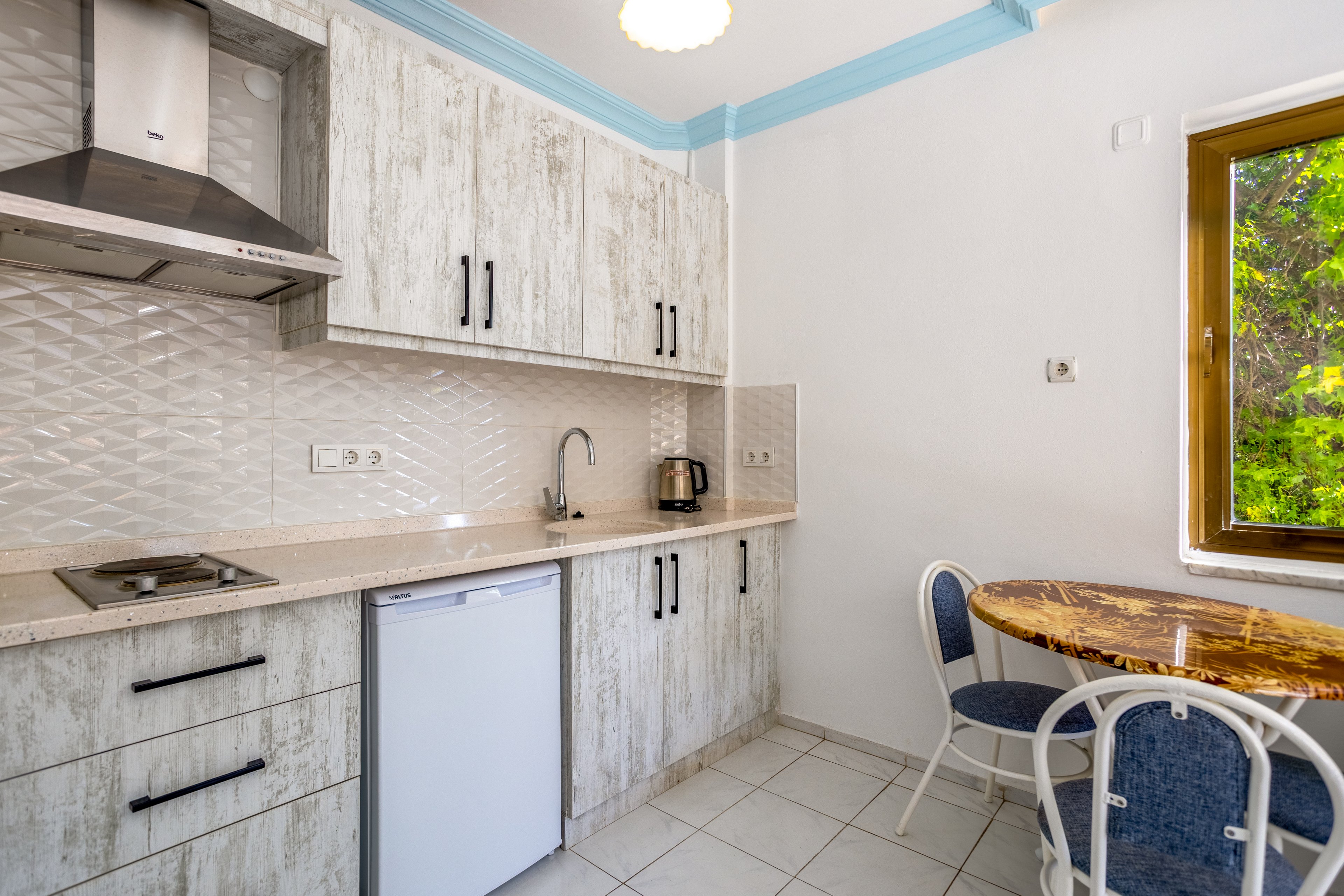 A compact modern kitchen features light wood cabinets, a small under-counter refrigerator, and a stainless steel range hood. The countertop holds a sink, electric cooktop, and a kettle, with textured white backsplash tiles adding visual interest. To the right, a round wooden table with blue and white chairs sits near a window overlooking green foliage. The space is clean, bright, and simply furnished, suggesting a cozy apartment or vacation rental kitchen.