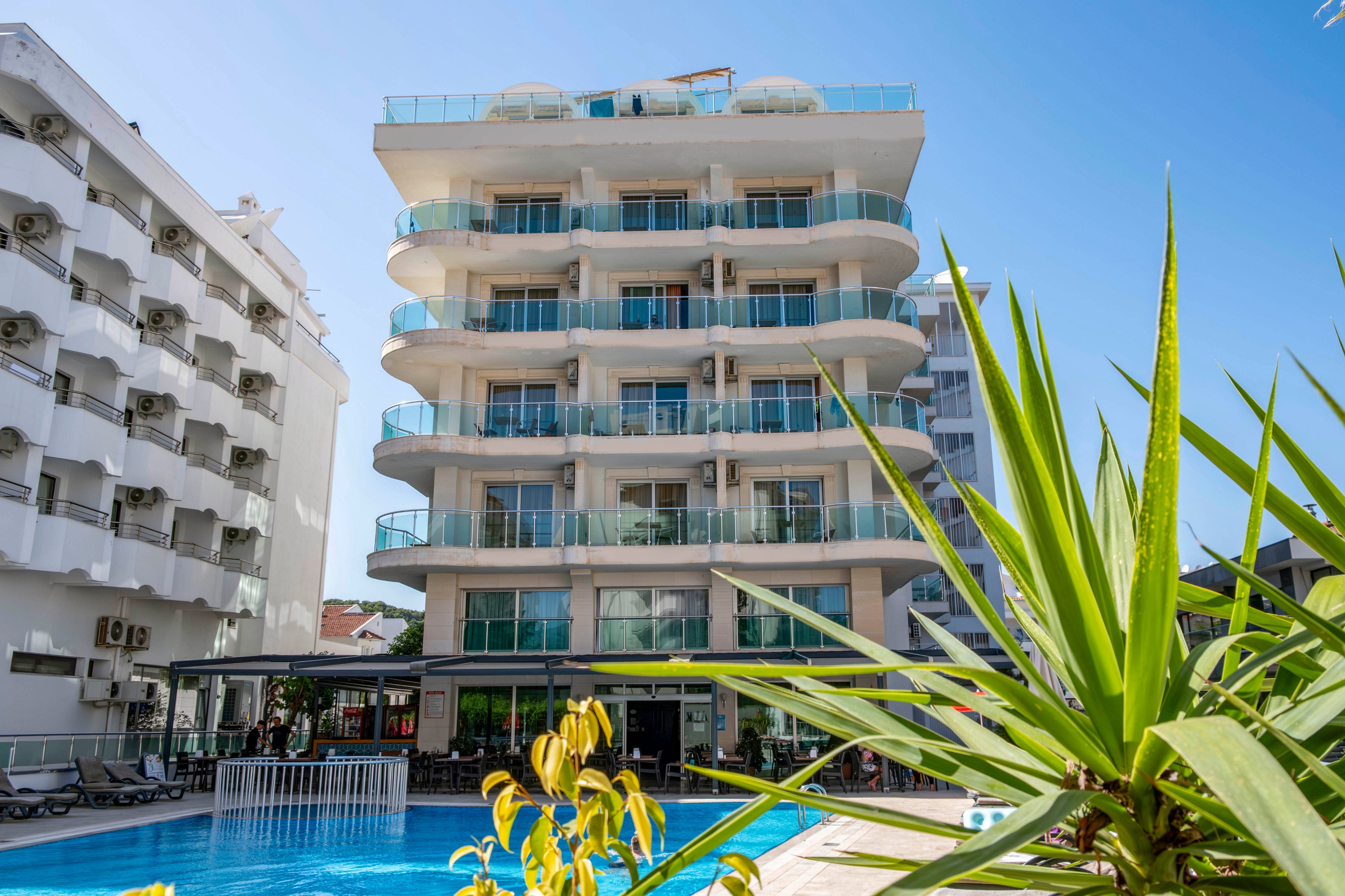 A mid-rise modern hotel stands prominently with glass balconies facing an outdoor swimming pool. The building features multiple floors with large windows and a clean, contemporary facade. Sun loungers and shaded seating areas surround the pool, suggesting a relaxing vacation setting. Neighboring buildings and clear blue sky frame the scene, while green plants add a touch of tropical atmosphere.