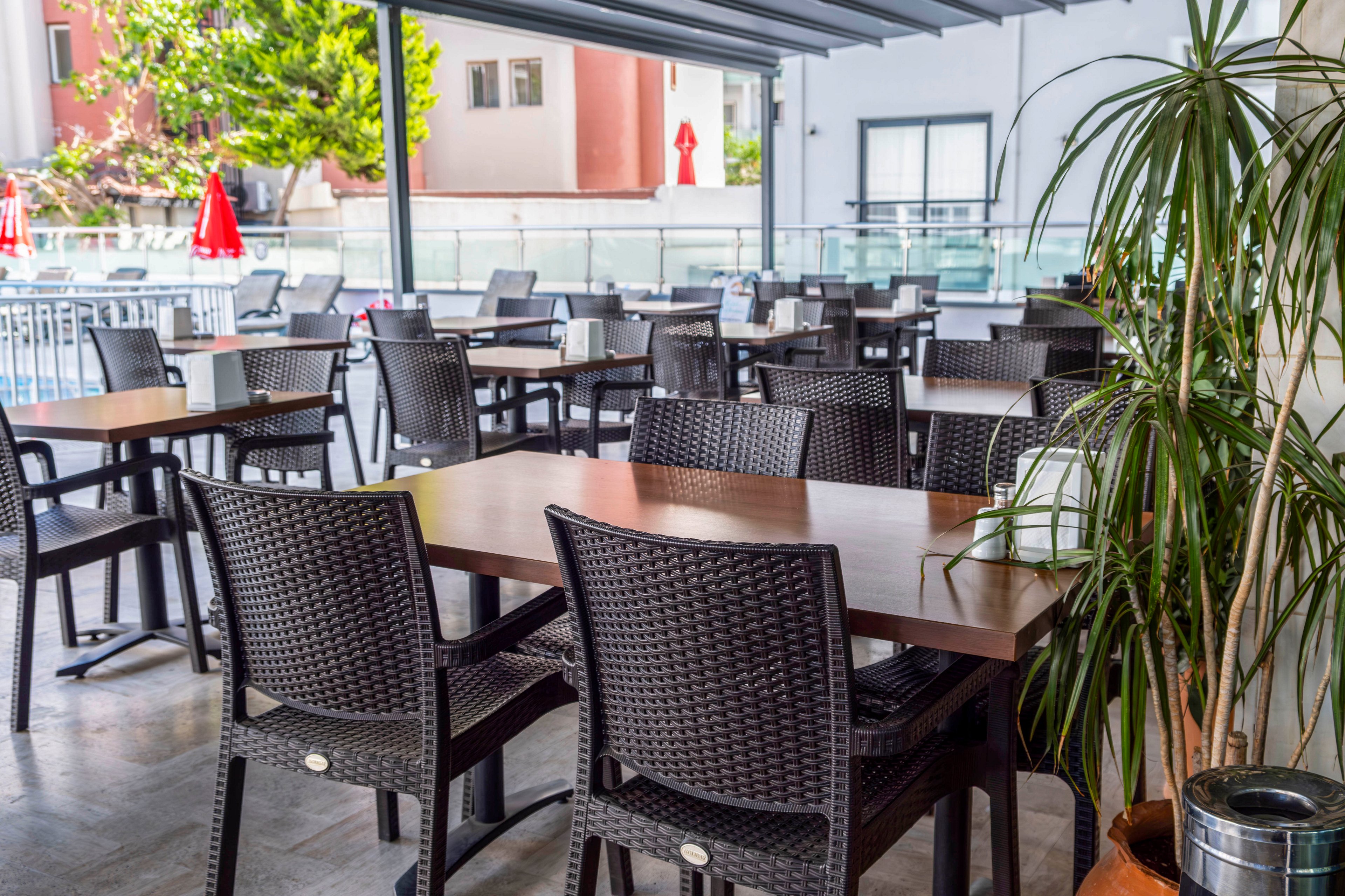 An empty outdoor cafe terrace features several wooden tables paired with dark wicker chairs. The seating area is covered by a modern metal canopy, providing shade and shelter. In the background, a glass railing separates the terrace from a pool or courtyard area with red umbrellas and nearby buildings. A tall potted plant adds greenery and a relaxed atmosphere to the scene.