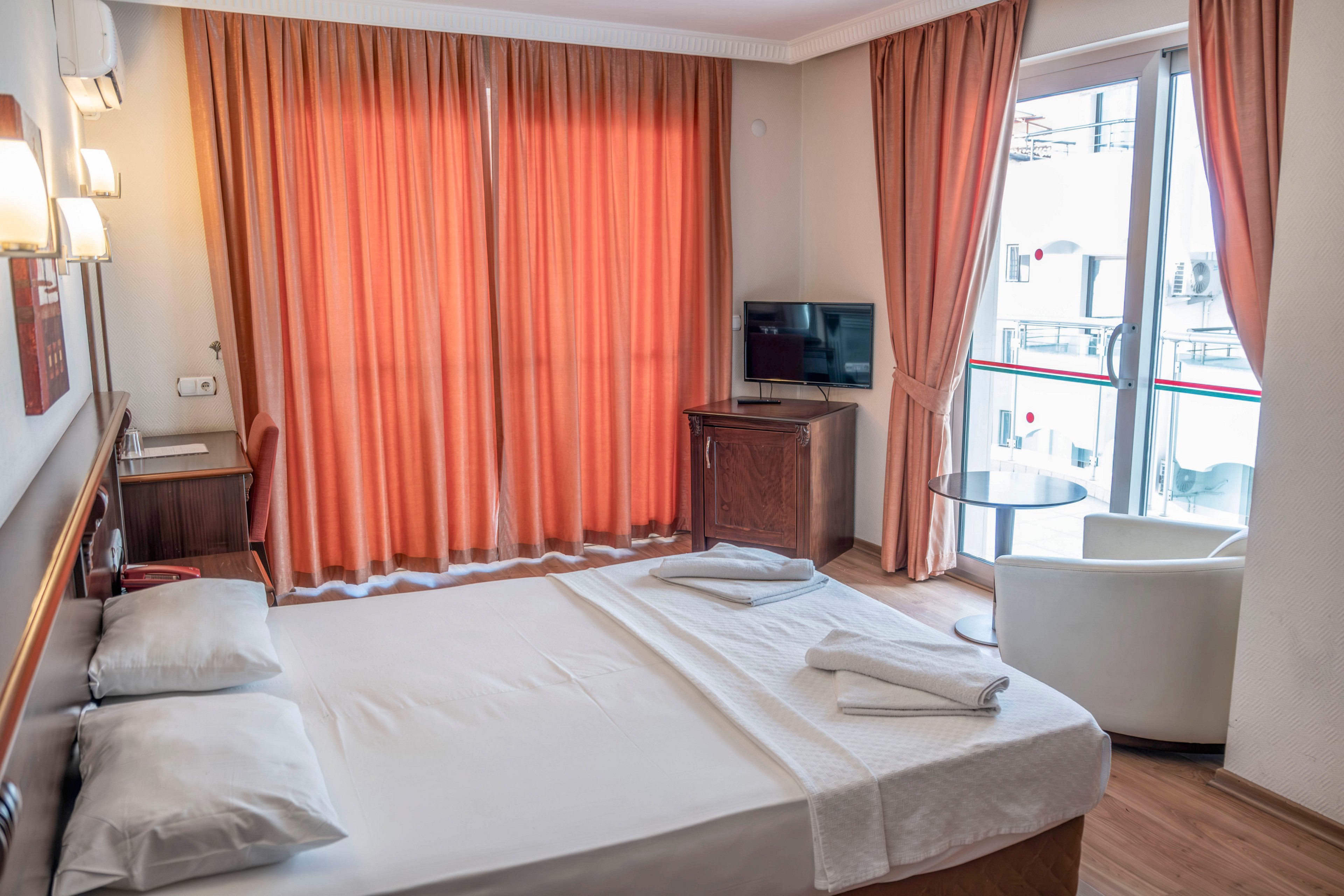 A neatly made double bed dominates a bright hotel room with white linens and folded towels. Warm orange curtains cover a large window, creating a cozy atmosphere. A small flat-screen television sits on a wooden cabinet opposite the bed. Near the balcony door, a round glass table and white armchair provide a small seating area with natural light.