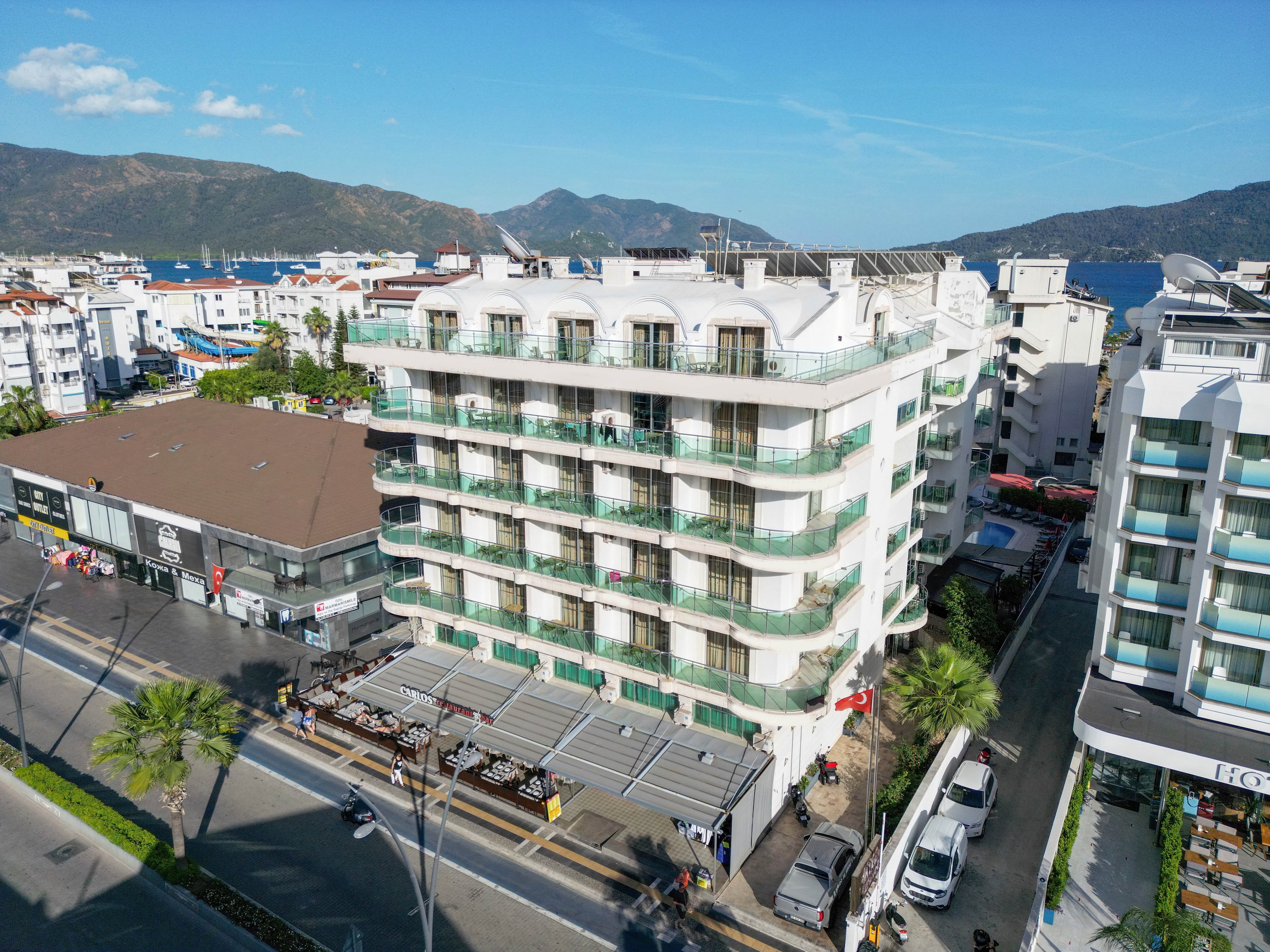 A multi-story modern hotel stands prominently along a coastal city street. The building features glass balconies on each floor and a ground-level terrace with outdoor seating. Surrounding it are other commercial buildings, palm-lined sidewalks, and parked cars. In the background, a calm sea and distant mountains frame the urban seaside setting under a clear blue sky.
