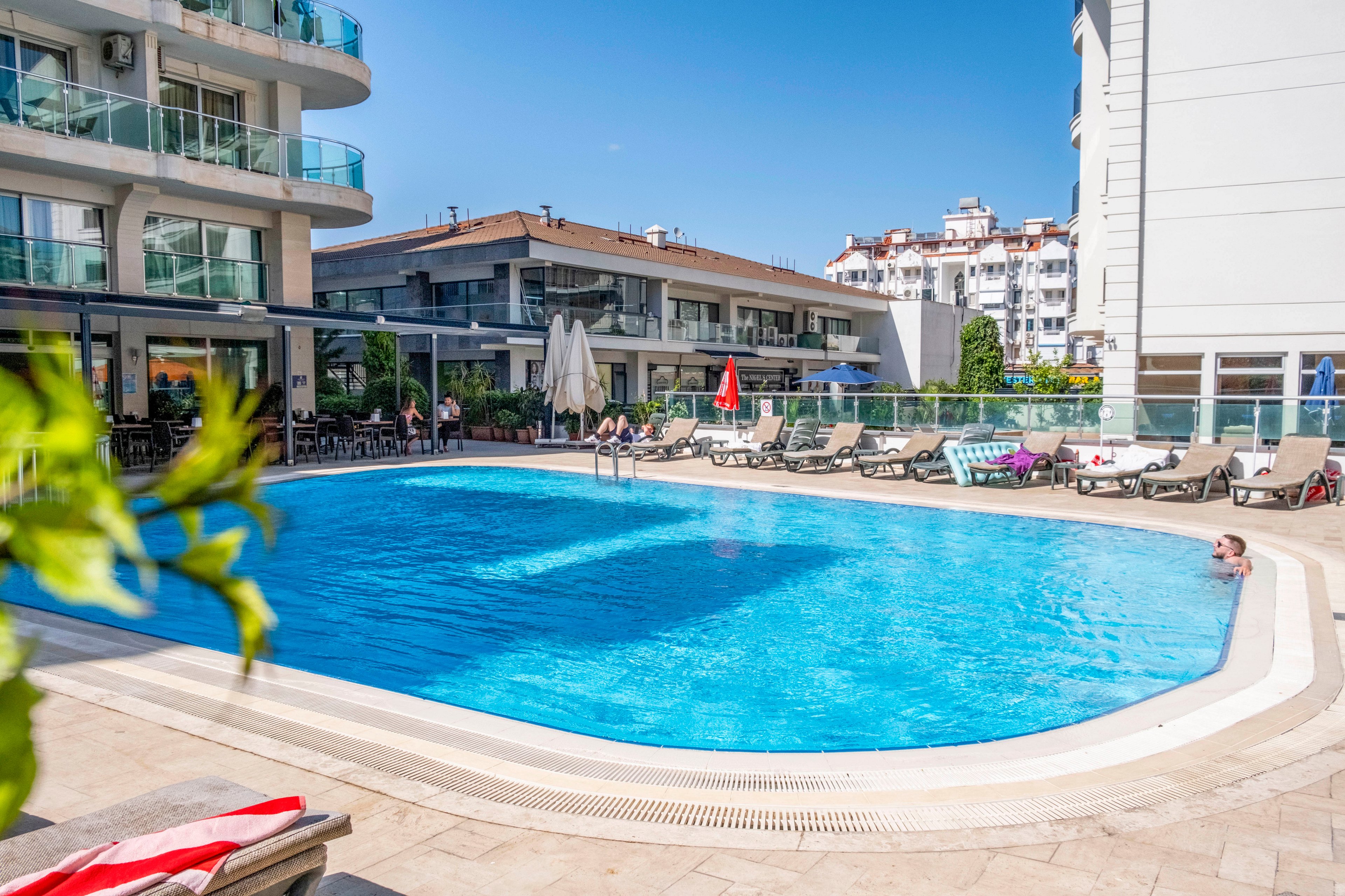 A large outdoor swimming pool sits at the center of a modern hotel courtyard. Sun loungers and umbrellas line the pool deck, with a few guests relaxing by the water. Contemporary multi-story buildings with balconies surround the pool area, suggesting an urban resort setting. Bright daylight and clear blue water create a clean, inviting vacation atmosphere.