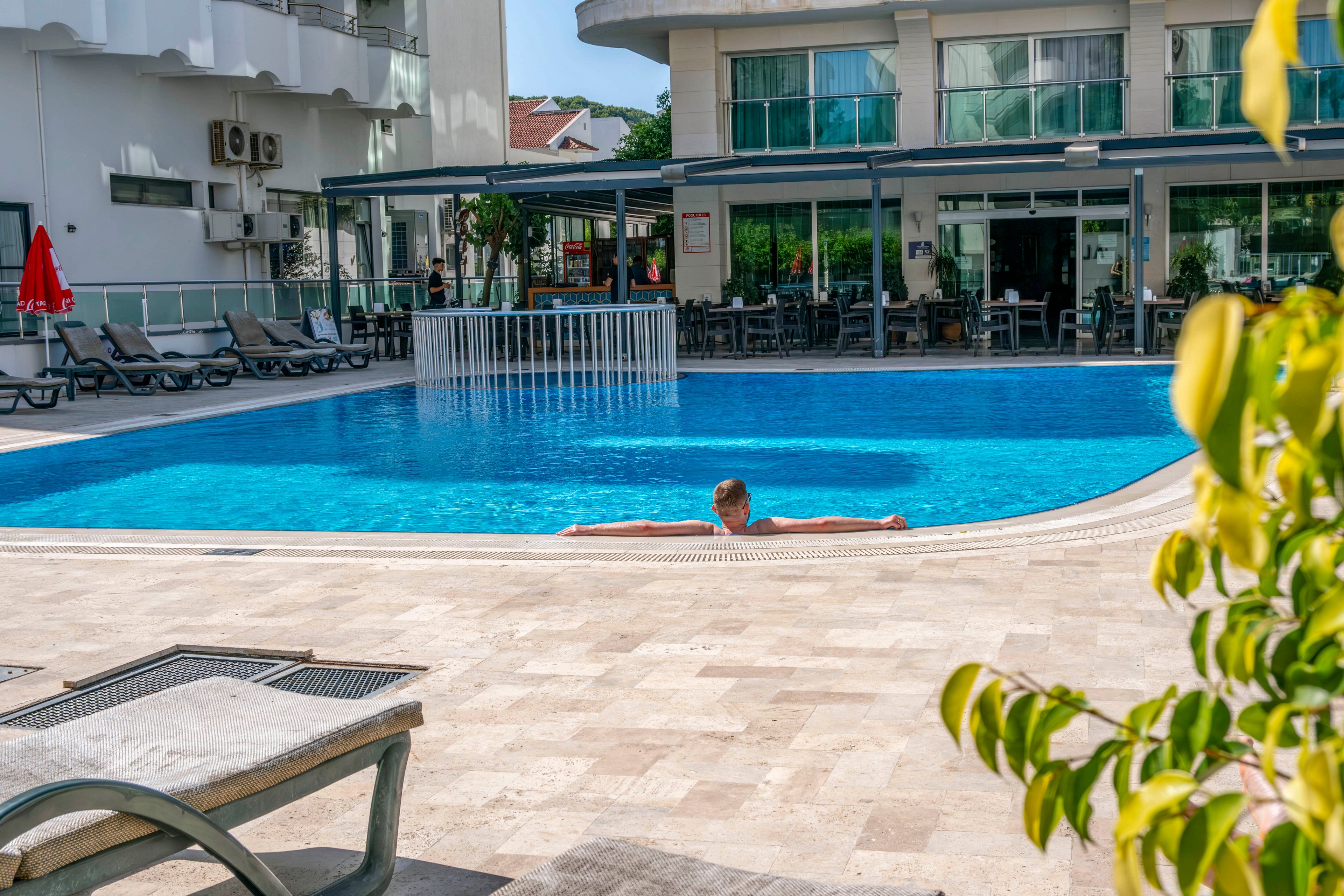 A person rests at the edge of a clear blue swimming pool in a modern hotel courtyard. Sun loungers and umbrellas line the poolside, suggesting a resort or vacation setting. The building’s glass windows and outdoor seating area frame the background, indicating a comfortable hospitality environment. Bright daylight and clean architectural lines create a calm, leisurely atmosphere.