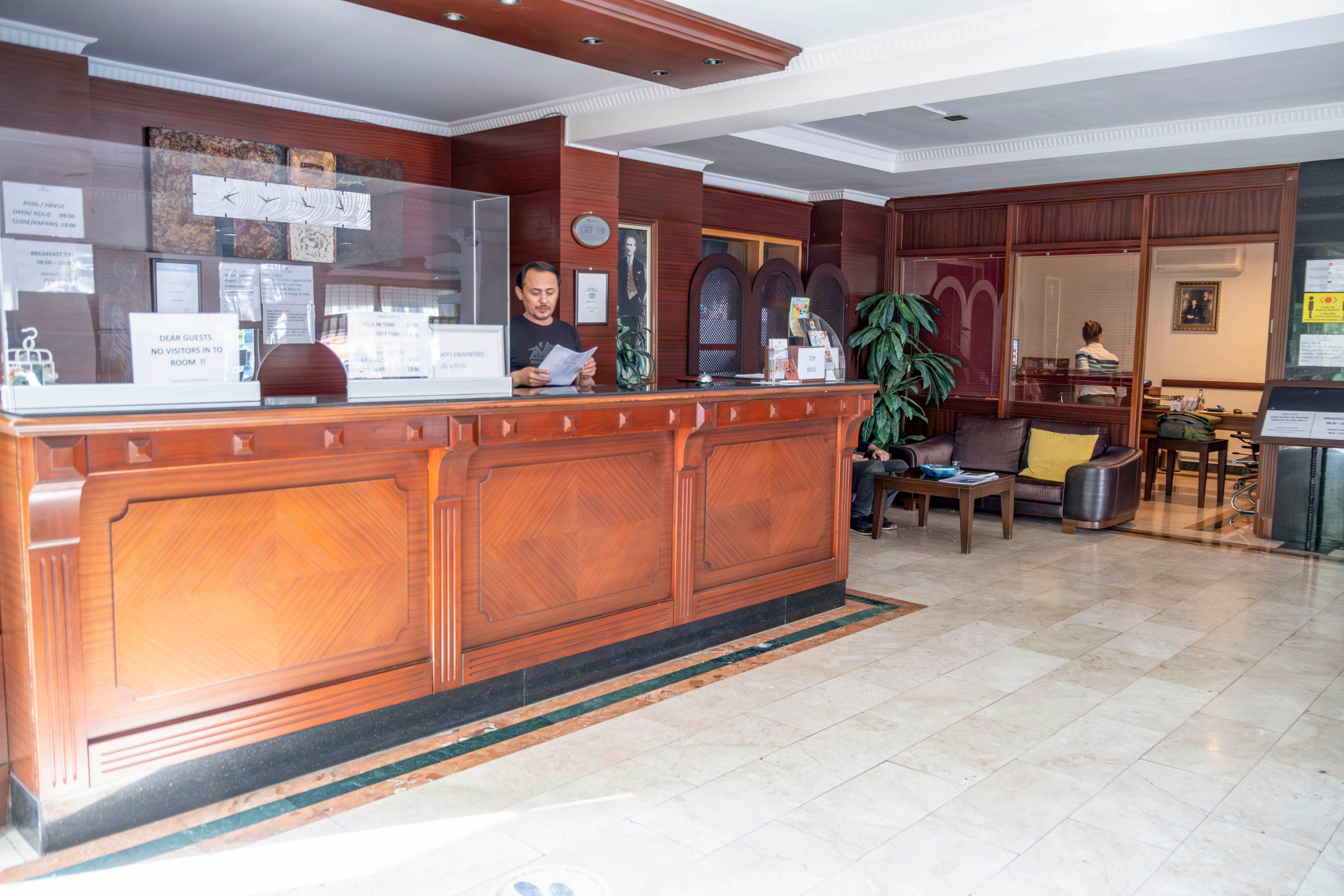 A hotel receptionist stands behind a large wooden front desk, reading documents in a bright lobby. The reception counter features classic wood paneling and a transparent protective screen. In the background, guests sit in a lounge area with armchairs and tables, and potted plants add greenery to the space. The marble floor and warm lighting create a welcoming, professional atmosphere.