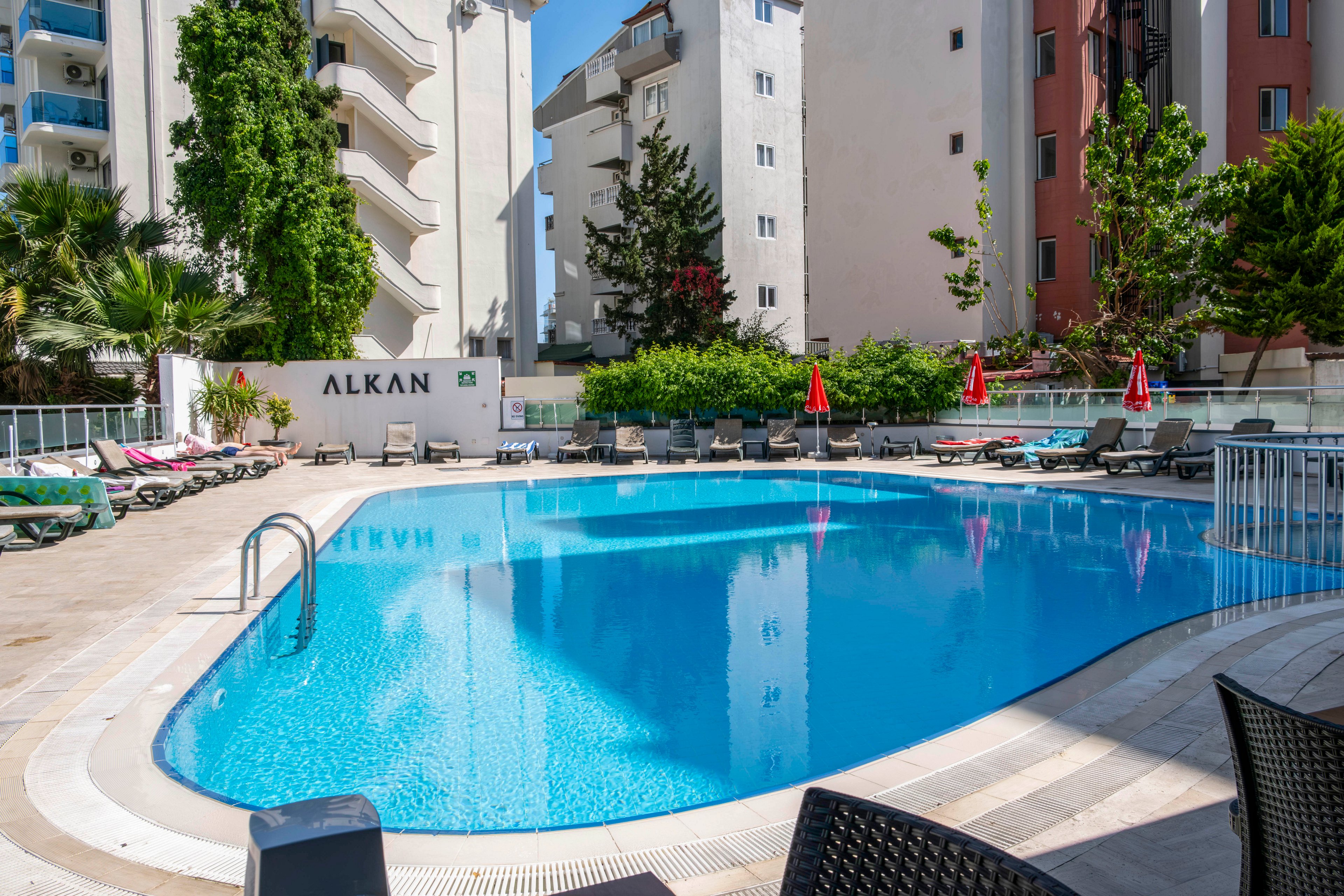 An outdoor hotel swimming pool is surrounded by sun loungers and umbrellas on a bright day. The clear blue water reflects nearby buildings and greenery, creating a relaxing vacation atmosphere. Apartment-style buildings and trees frame the pool area, suggesting an urban resort setting. The word "ALKAN" is visible on a wall near the pool, indicating the property name.