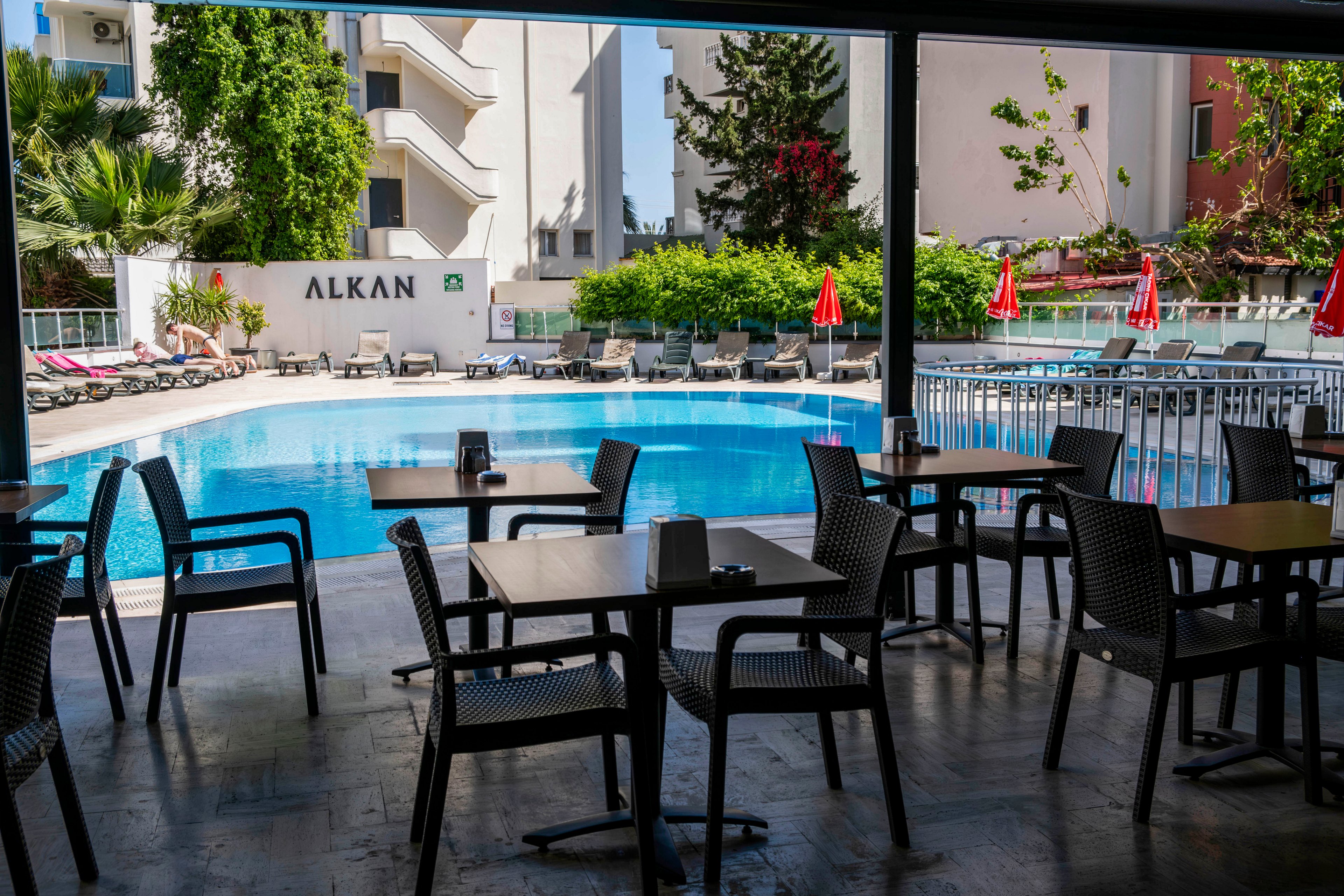An outdoor hotel swimming pool is viewed from a shaded cafe seating area. Empty tables and chairs are arranged in the foreground under a covered terrace. Sun loungers and red umbrellas line the poolside, suggesting a relaxed vacation setting. A white building and lush green trees surround the pool, creating a bright and inviting atmosphere. The word "ALKAN" is visible on the building near the pool.